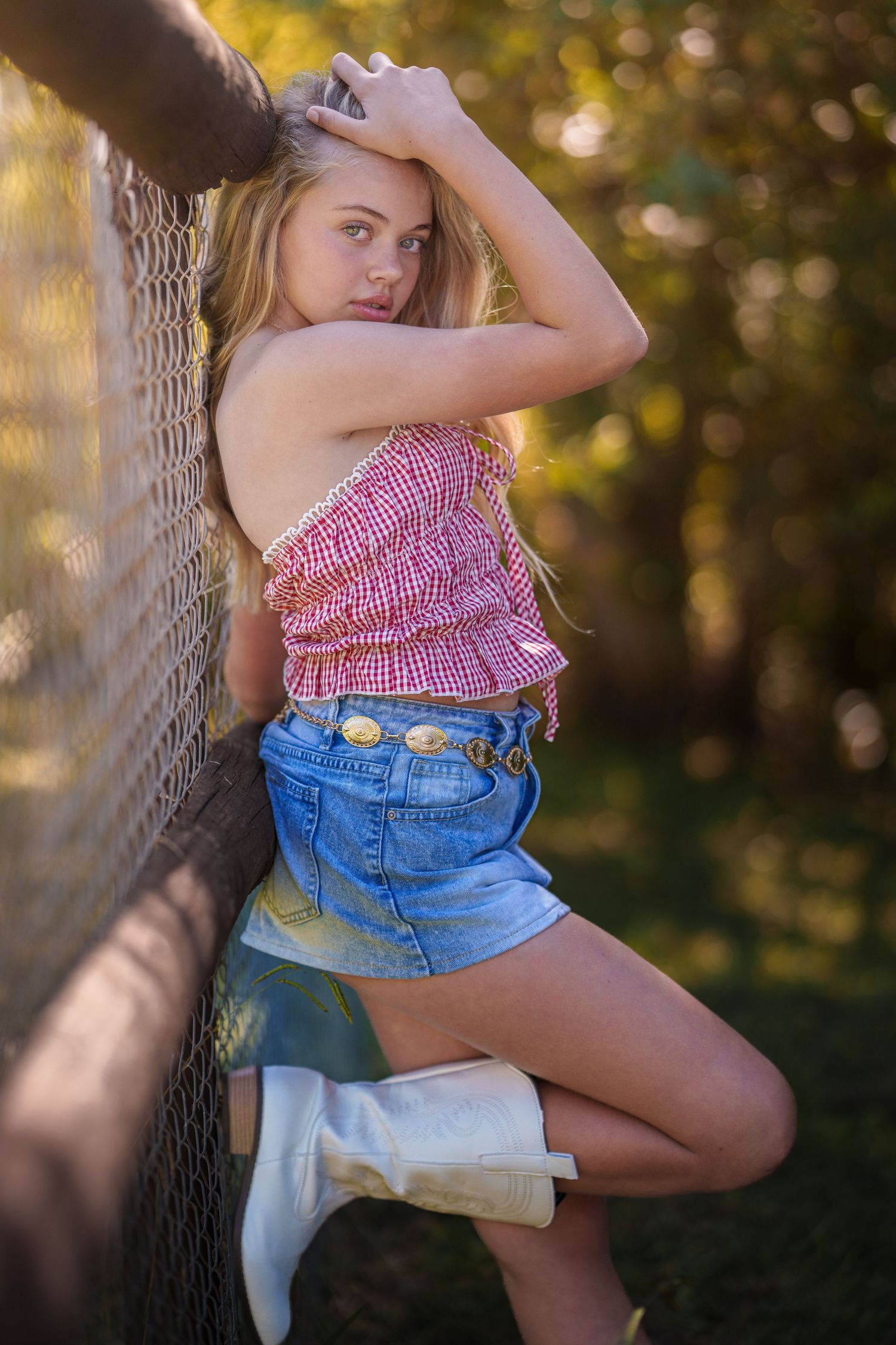 Young woman in red gingham top, denim shorts, and white cowboy boots posing against a fence outdoors.