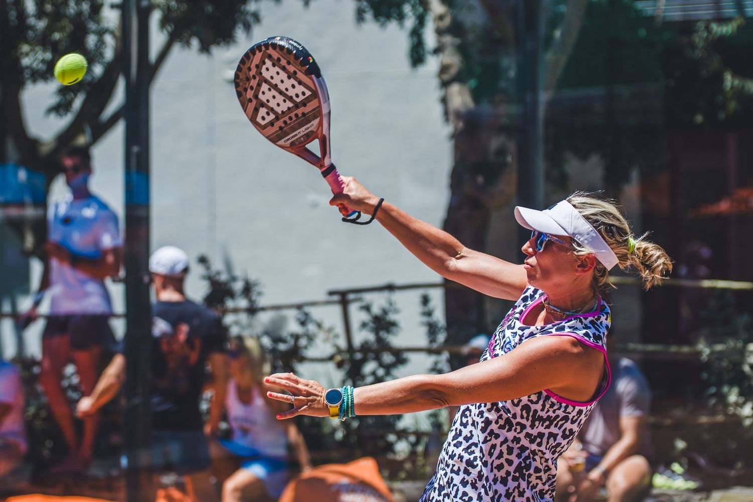 Woman in floral outfit playing padel tennis, swinging racket at yellow ball on outdoor court in sunny conditions.