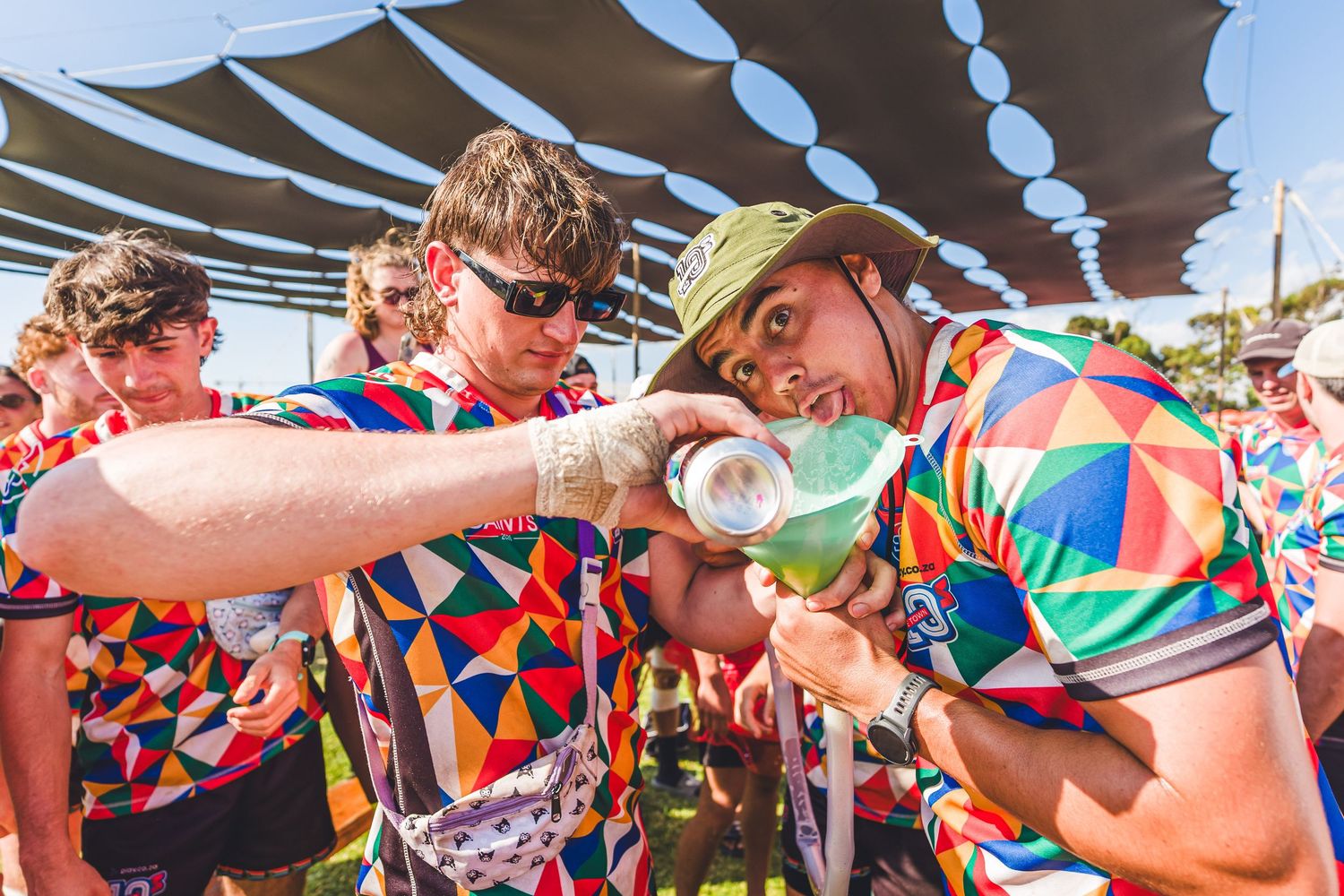Two cyclists in colorful geometric jerseys share a water bottle at an outdoor cycling event on a sunny day.