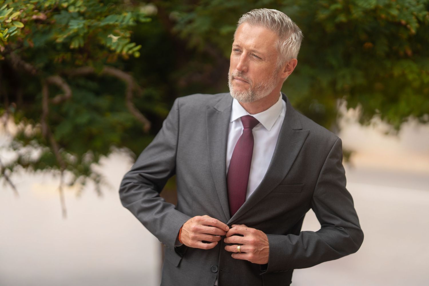 Man in grey suit and burgundy tie posing outdoors near autumn foliage.