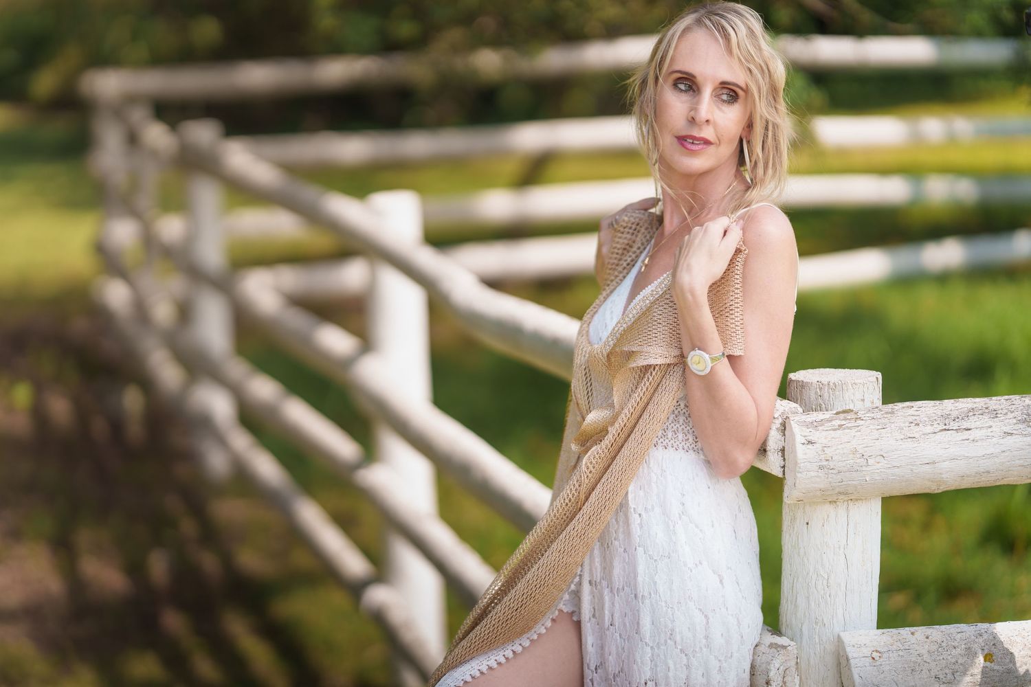 A person in a white dress poses outdoors by a rustic wooden fence in a sunlit garden setting.