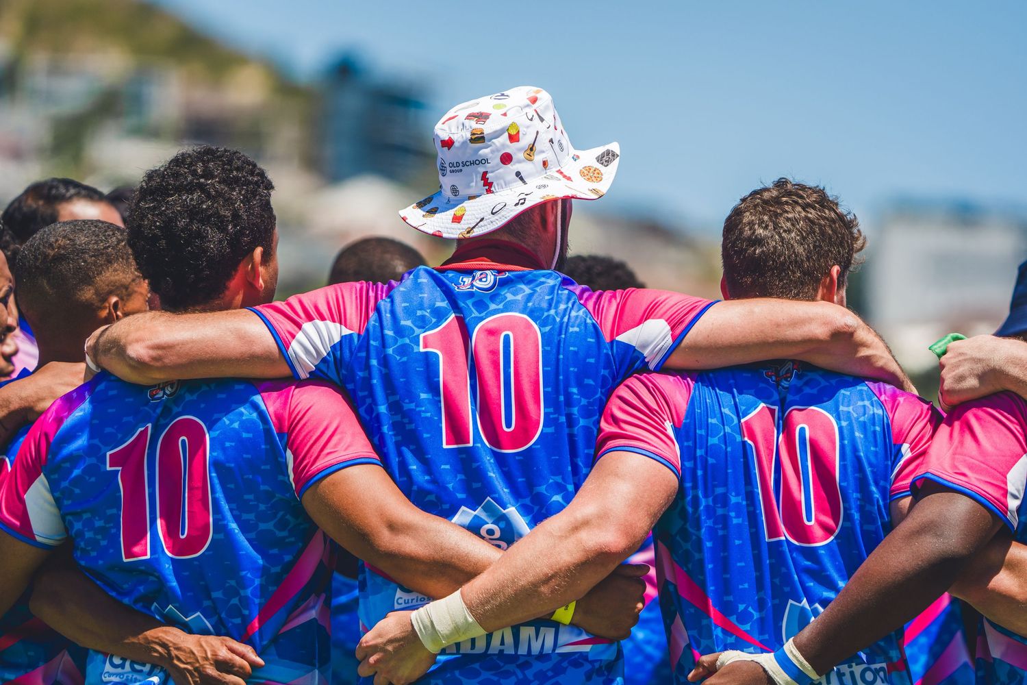 Rugby team huddle in blue and pink number 10 jerseys on a sunny beach, viewed from behind.