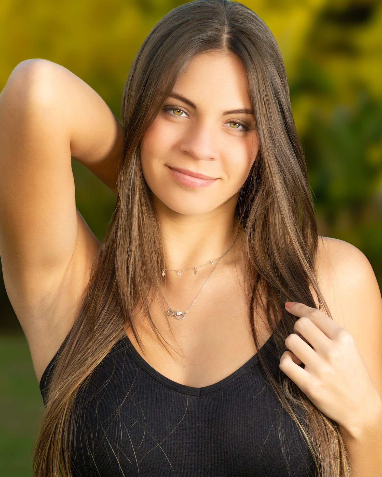 Young woman with long brown hair wearing a black tank top and silver necklace, smiling outdoors in natural light.