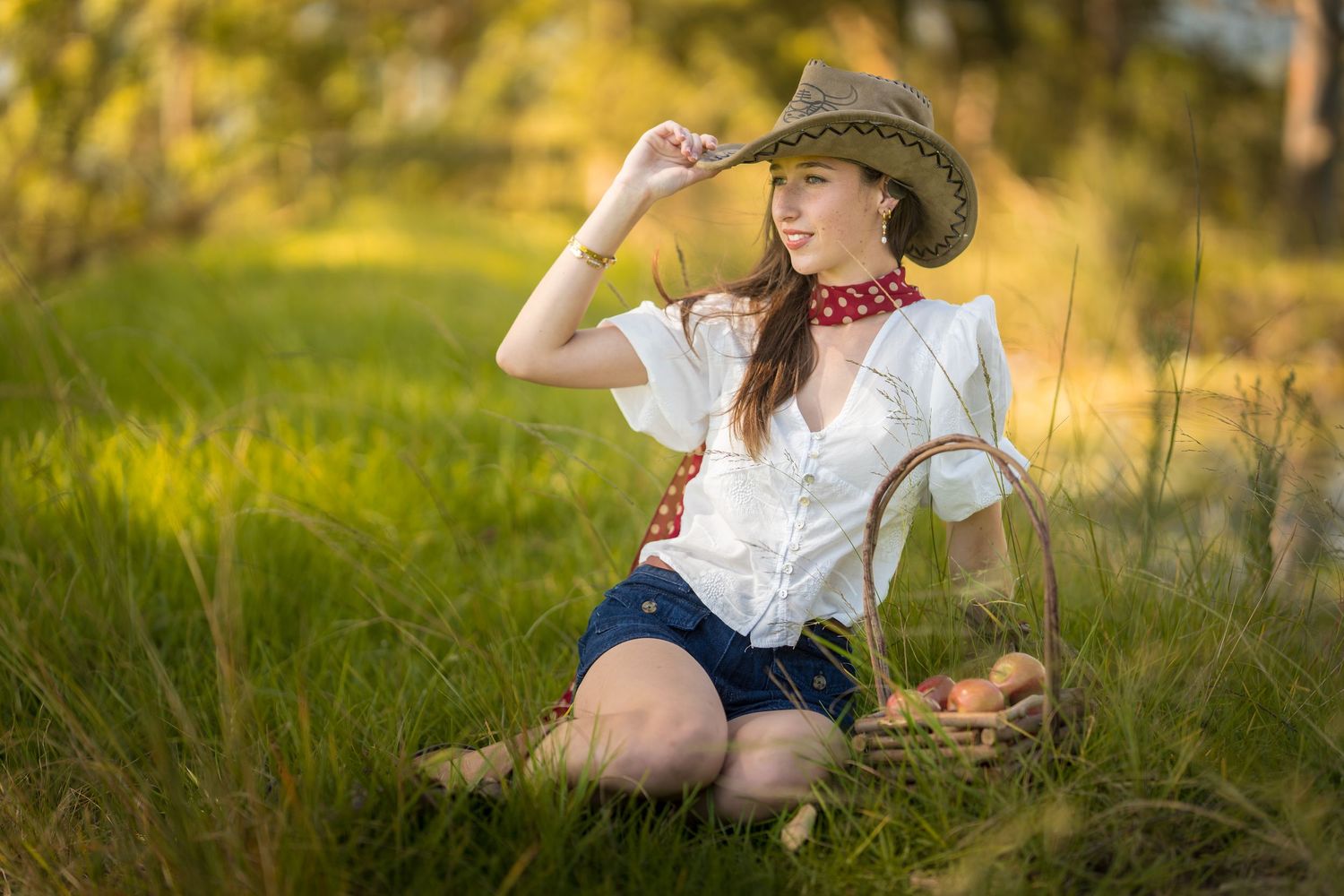 Young woman in cowboy hat and white blouse sits on grass with wicker basket in sunny outdoor setting.