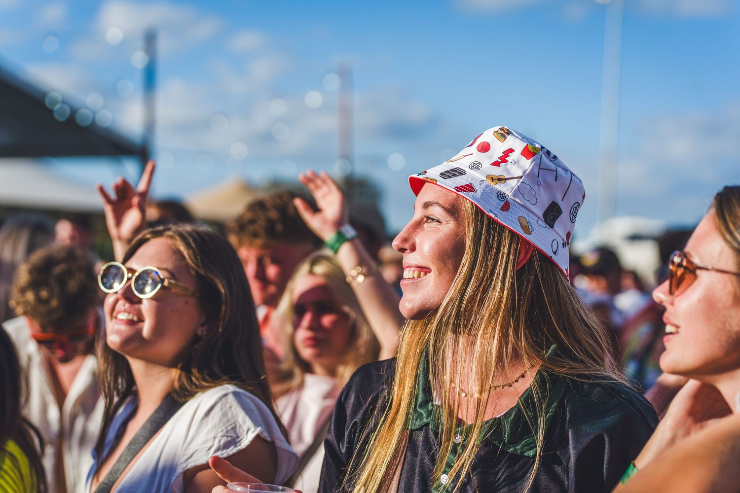 Young women smiling and enjoying a sunny outdoor music festival, one wearing a colorful bucket hat and white sunglasses.