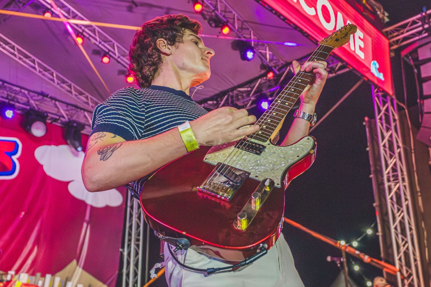 Guitarist performing on stage at night, playing a red electric guitar under colorful festival lights.