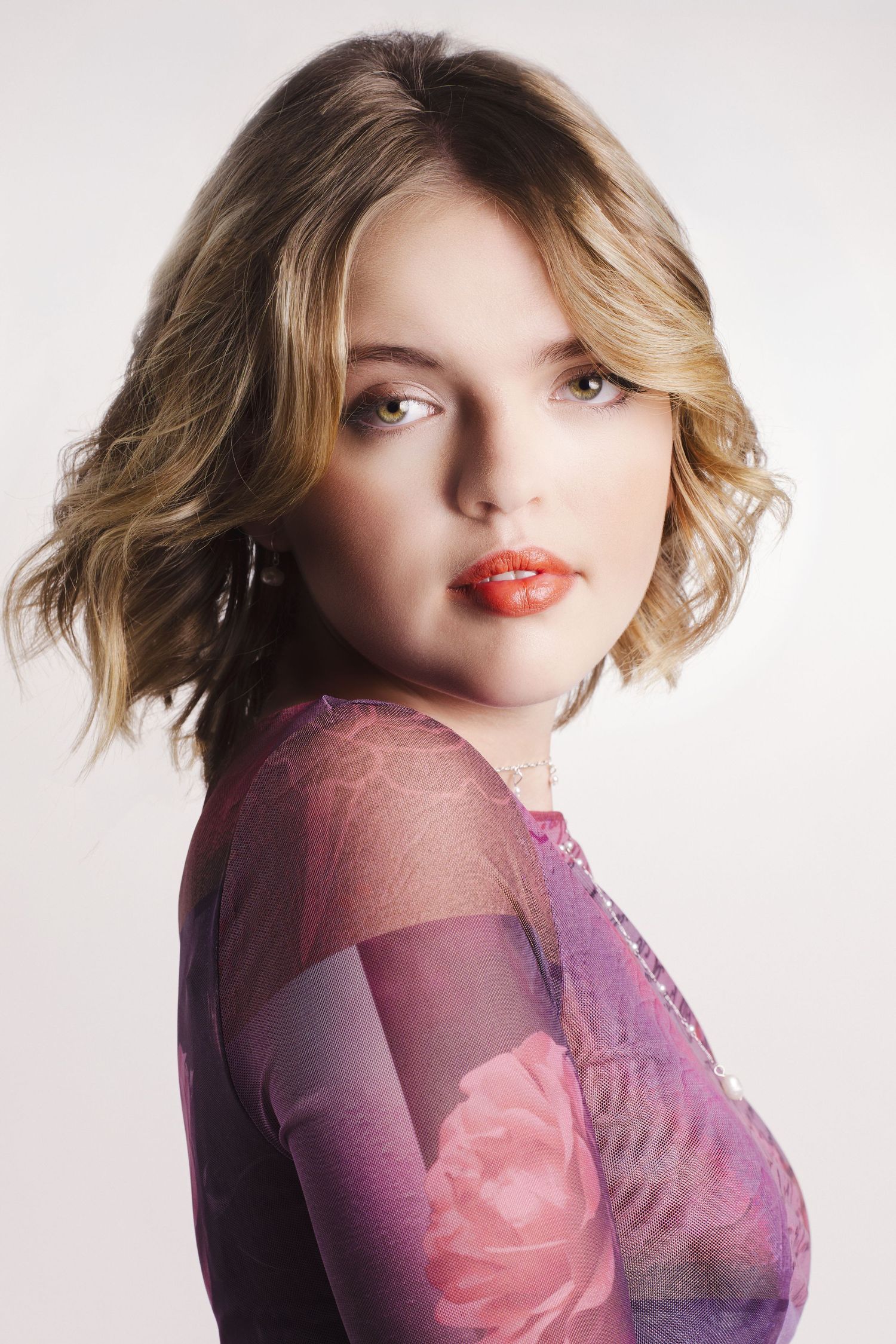 Young woman with wavy blonde hair and red lips wearing a purple floral lace top poses against white background.