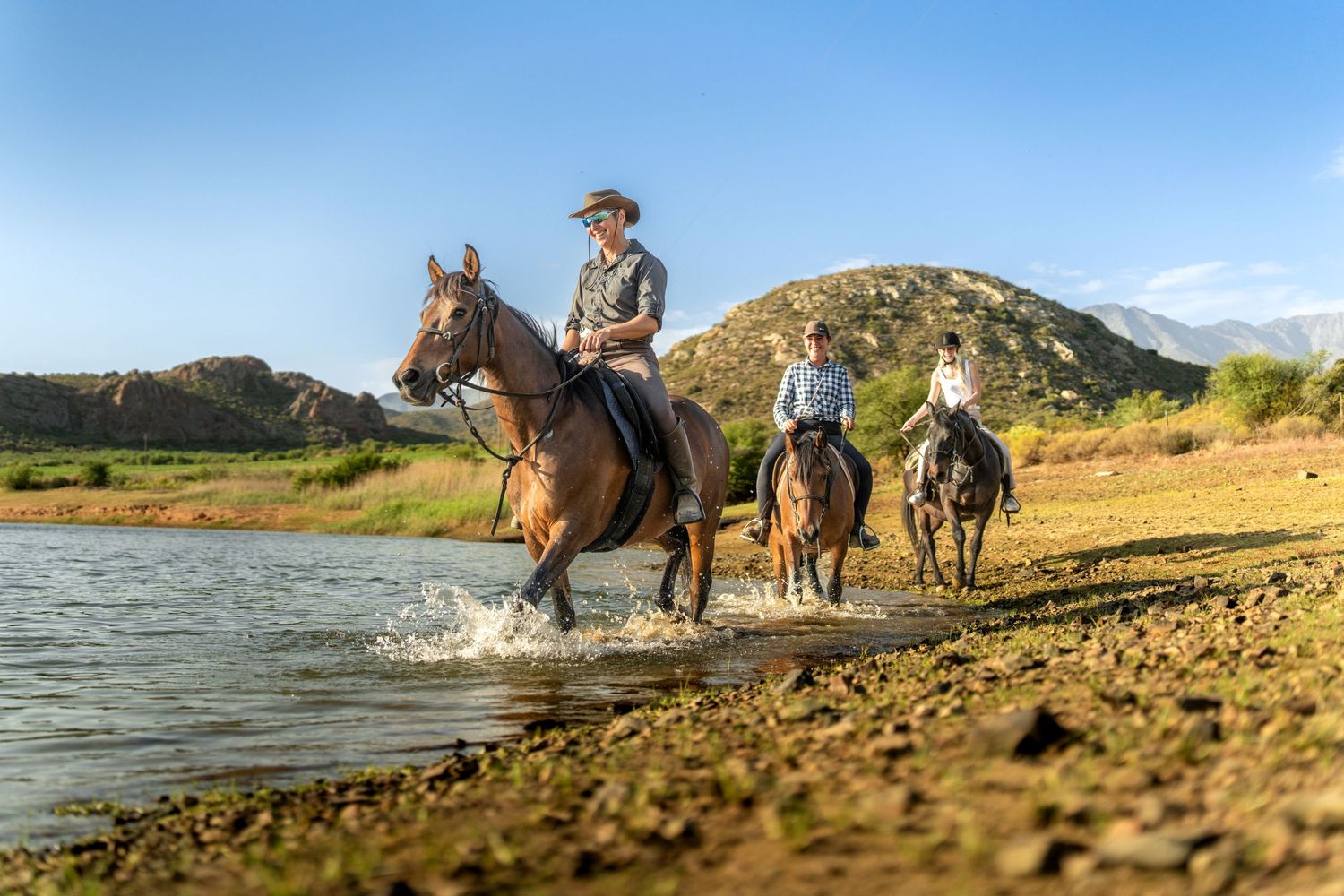 Three horseback riders riding through a pond with karoo views in the background