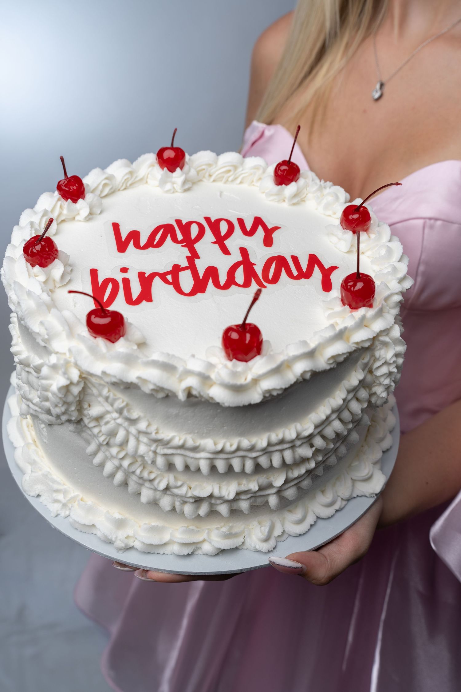 A white birthday cake decorated with whipped cream, cherries and red lettering.