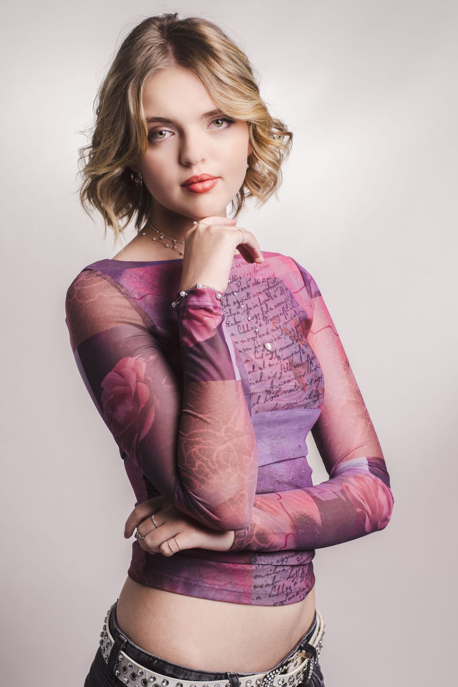 Young blonde woman in pink floral mesh long-sleeve top posing thoughtfully against light gray background.