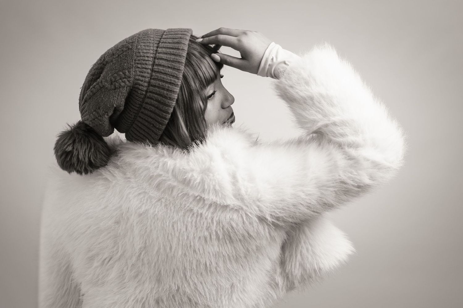 Black and white photo of a woman in a fluffy white coat and knit beanie hat posing against a light background.