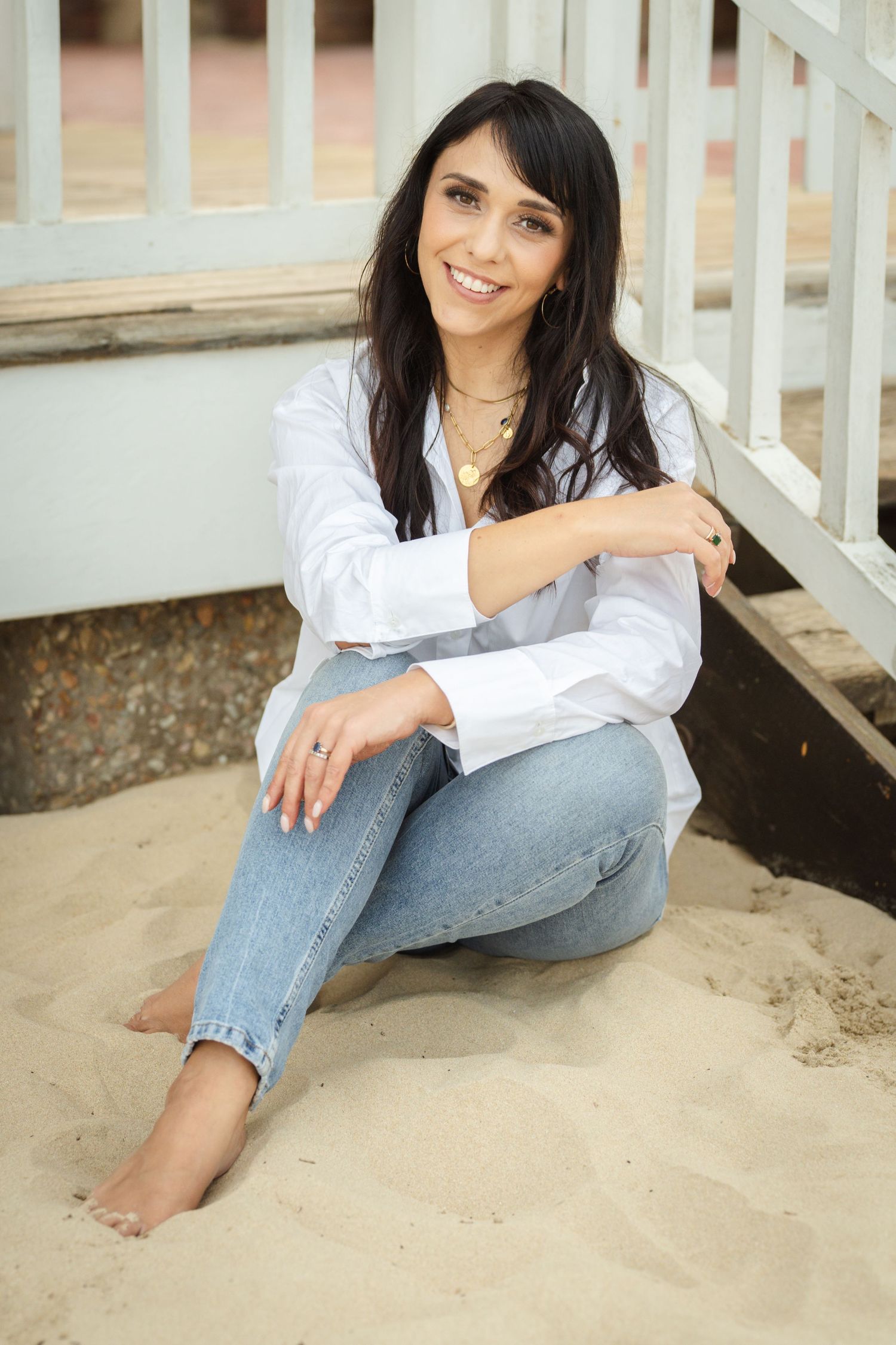 Smiling woman with dark hair sits on sandy beach steps, wearing white shirt and jeans, barefoot.