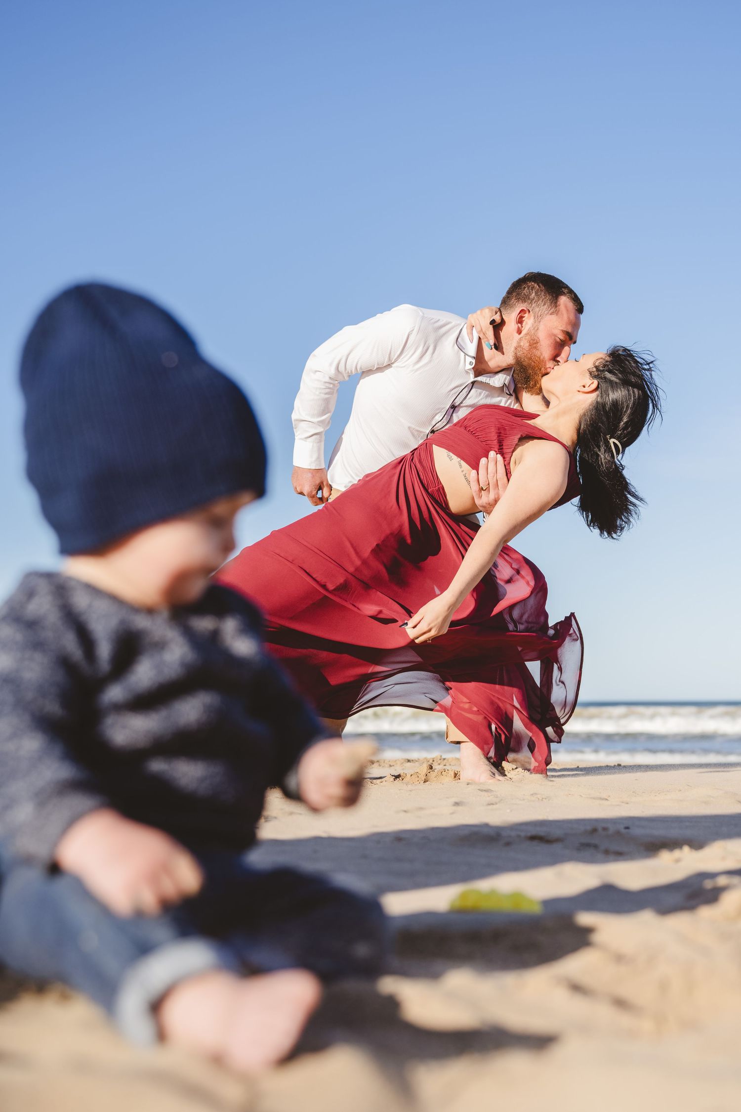 Romantic beach photoshoot with couple in red dress and white shirt against ocean backdrop.