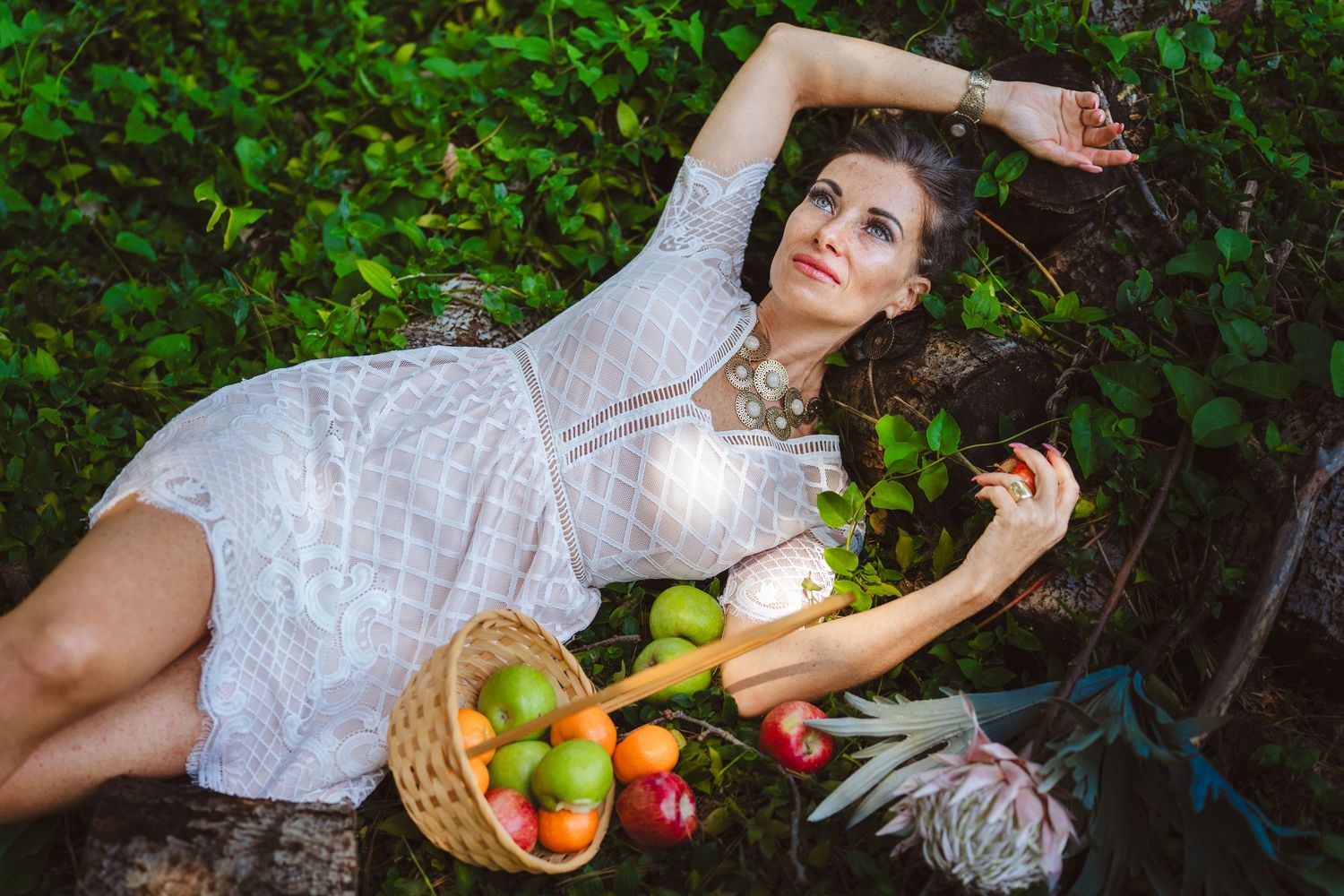A person in a white dress lies on grass surrounded by fresh apples and flowers in a dreamy garden setting.