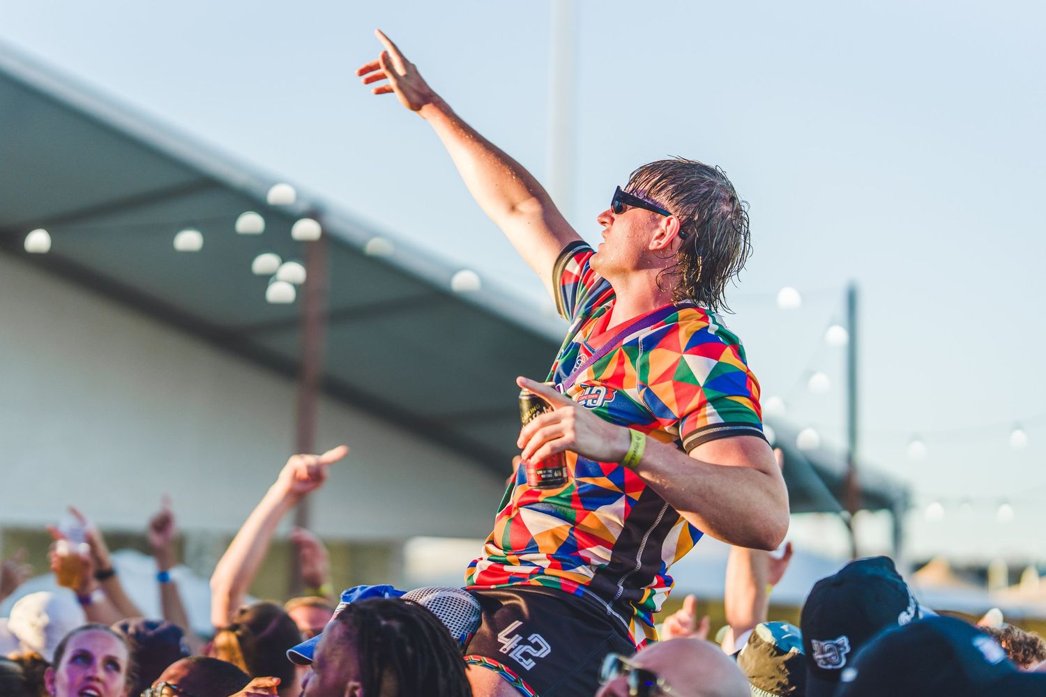 Excited festival-goer in colorful shirt crowd surfing and pointing skyward at an outdoor music festival.