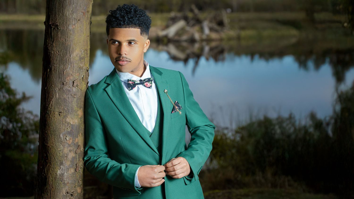 Young man in emerald green tuxedo and bow tie poses outdoors by a lake.