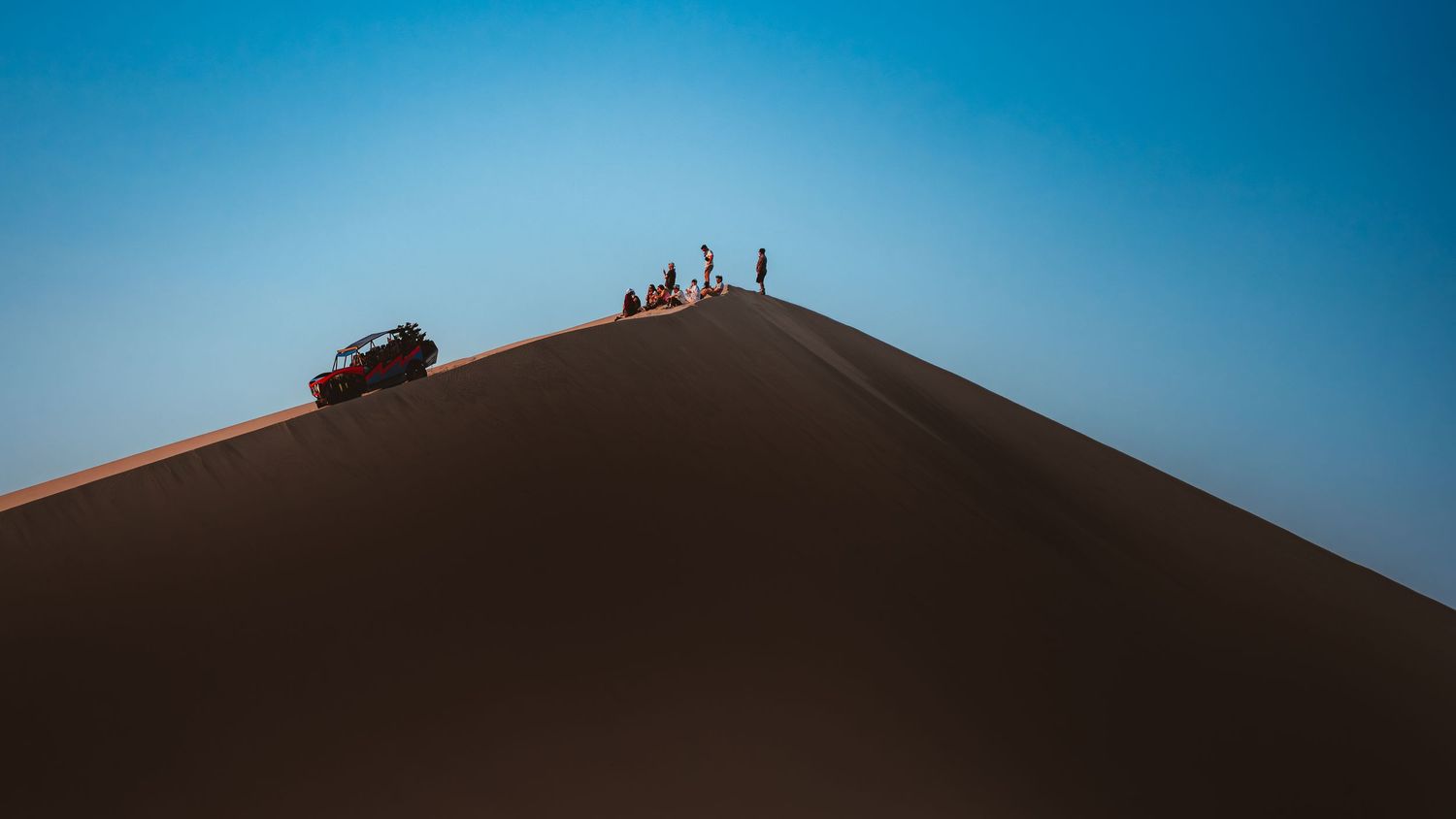 A tourist group watching the sunset on top of a sand dune peak with their dune buggy a bit lower down on the dune