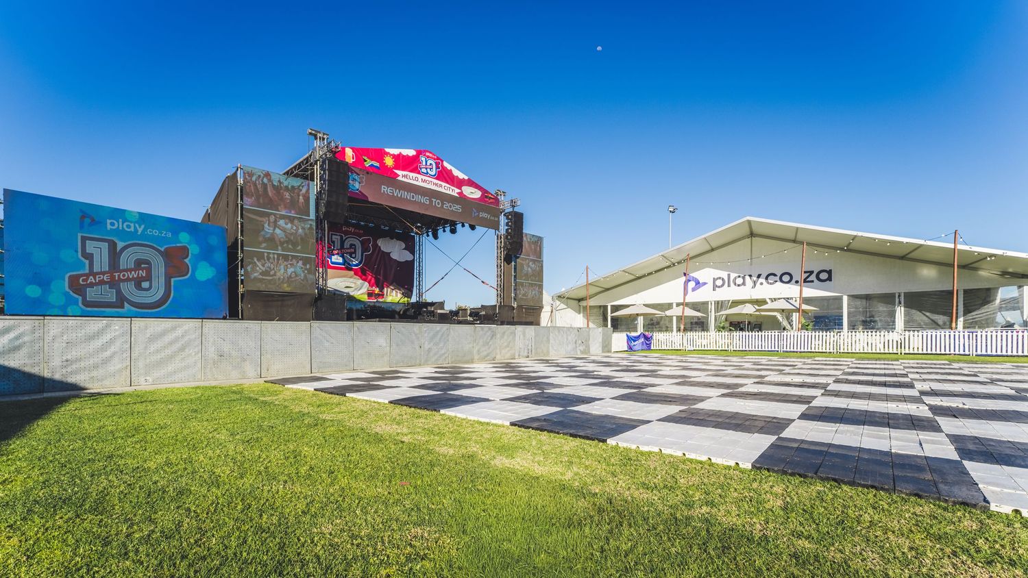 Outdoor concert stage with checkered floor pattern and green lawn at a racing venue under clear blue sky.