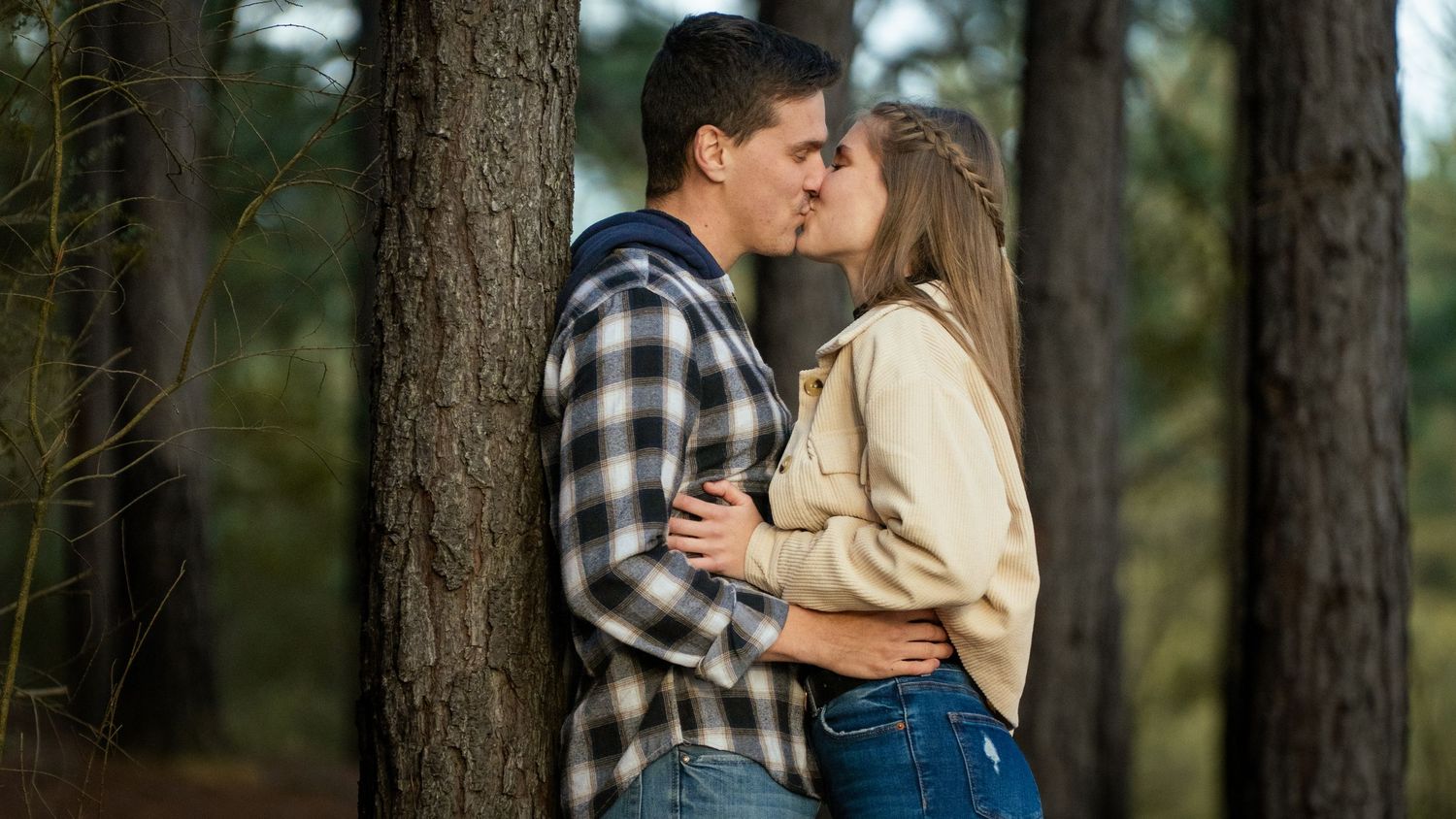A couple shares an intimate moment in a wooded forest setting with sunlight filtering through the trees.