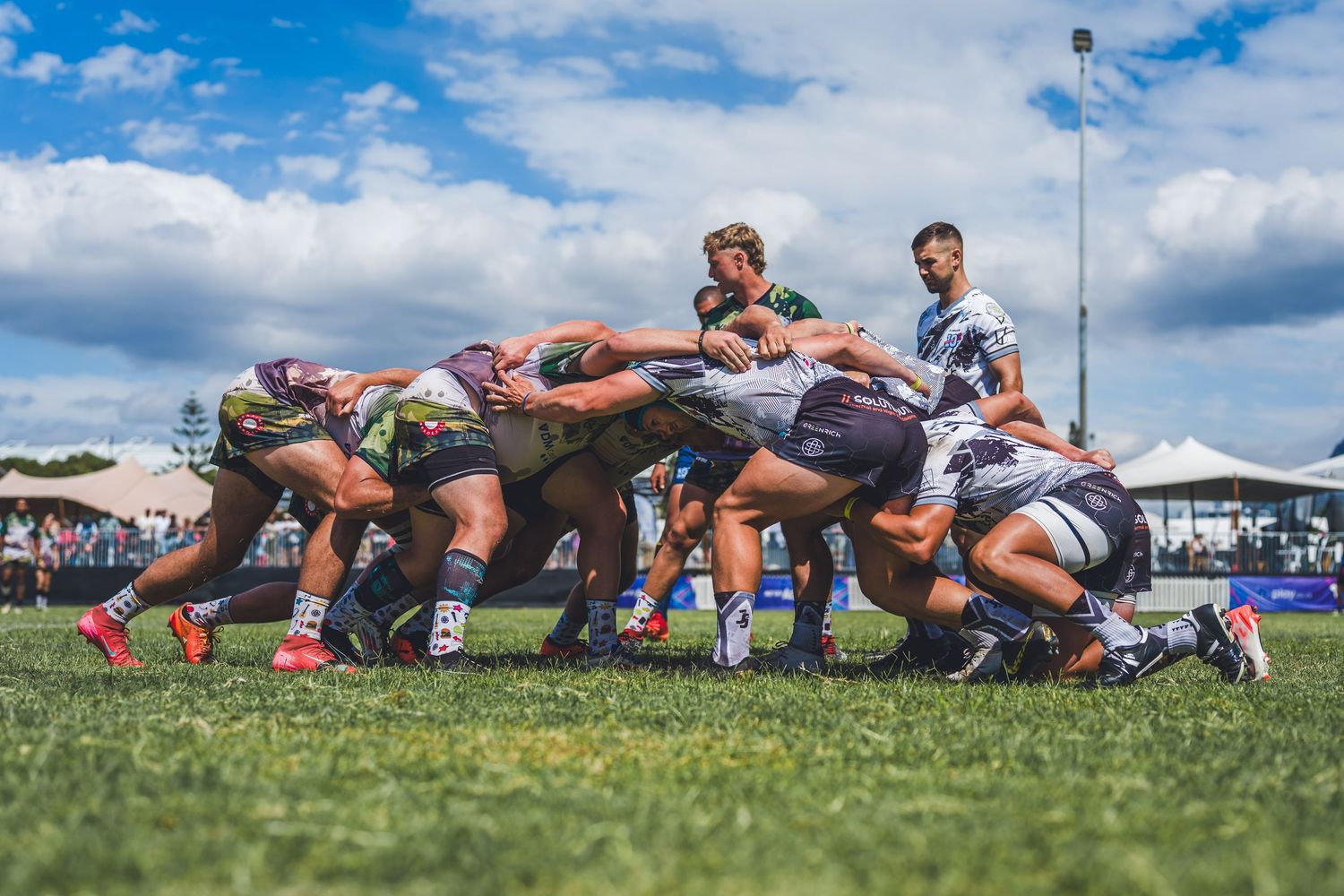 Rugby players engage in an intense scrum during a competitive match on a green field under a partly cloudy sky.