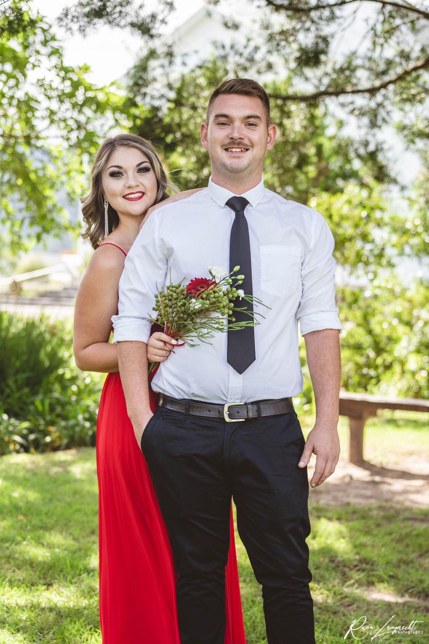 A couple poses together outdoors in formal attire with a red dress and white shirt with tie.