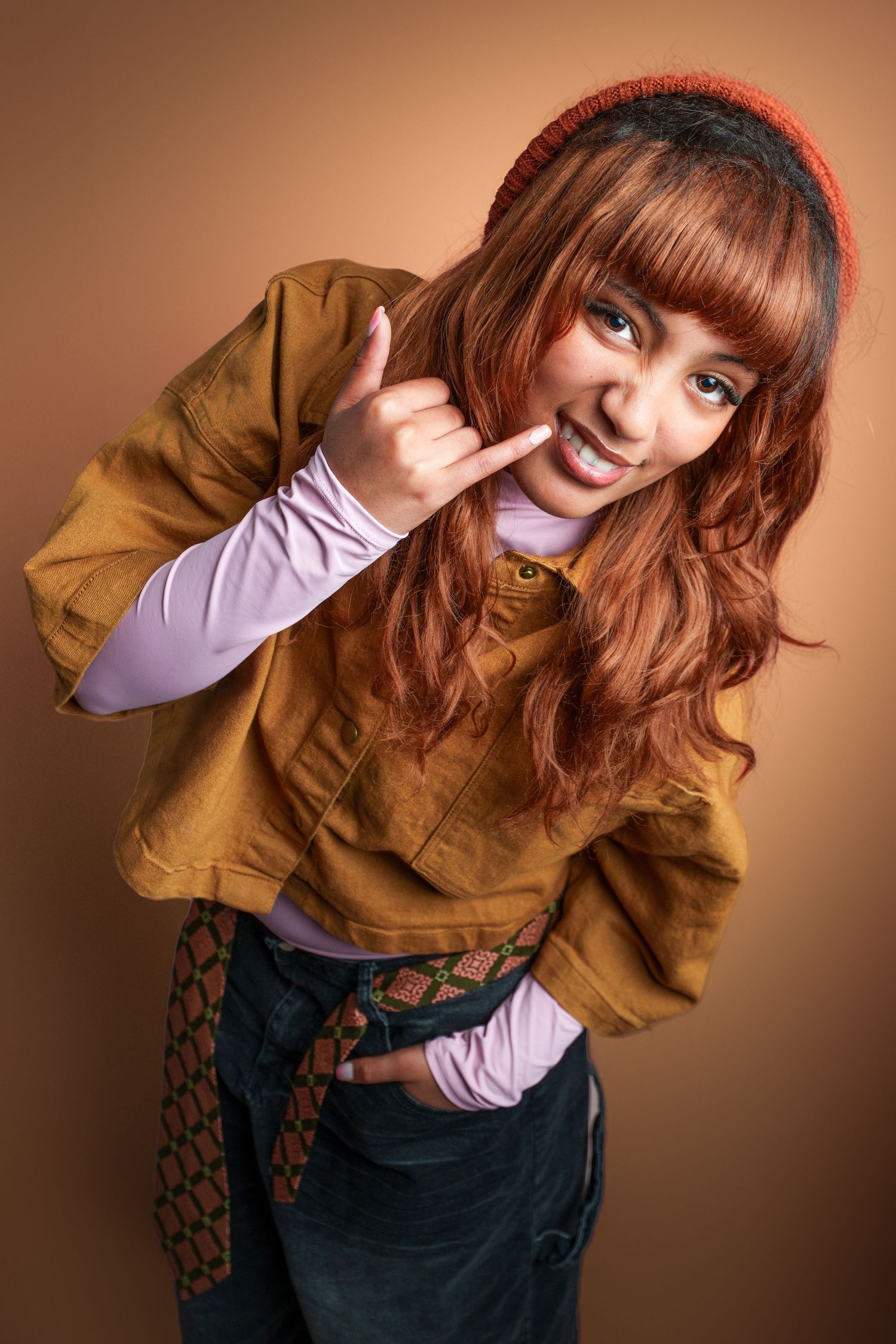 Young woman with red hair and beanie making a rock sign gesture, wearing brown jacket, smiling at camera.