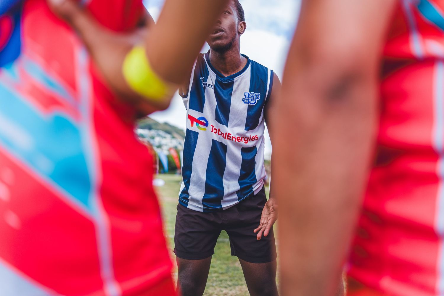 A soccer player in a blue and white striped jersey stands on a field surrounded by teammates in red uniforms.