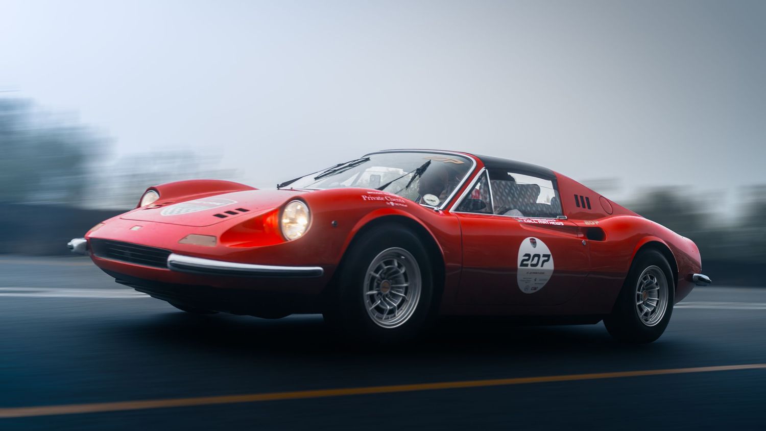 Red Ferrari Dino 246 GT classic sports car racing on a track, number 207, captured in motion with a blurred background.