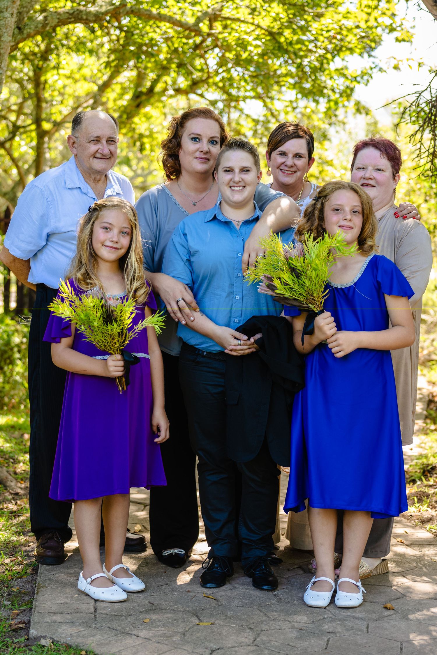 A multigenerational family group poses outdoors holding purple flowers.