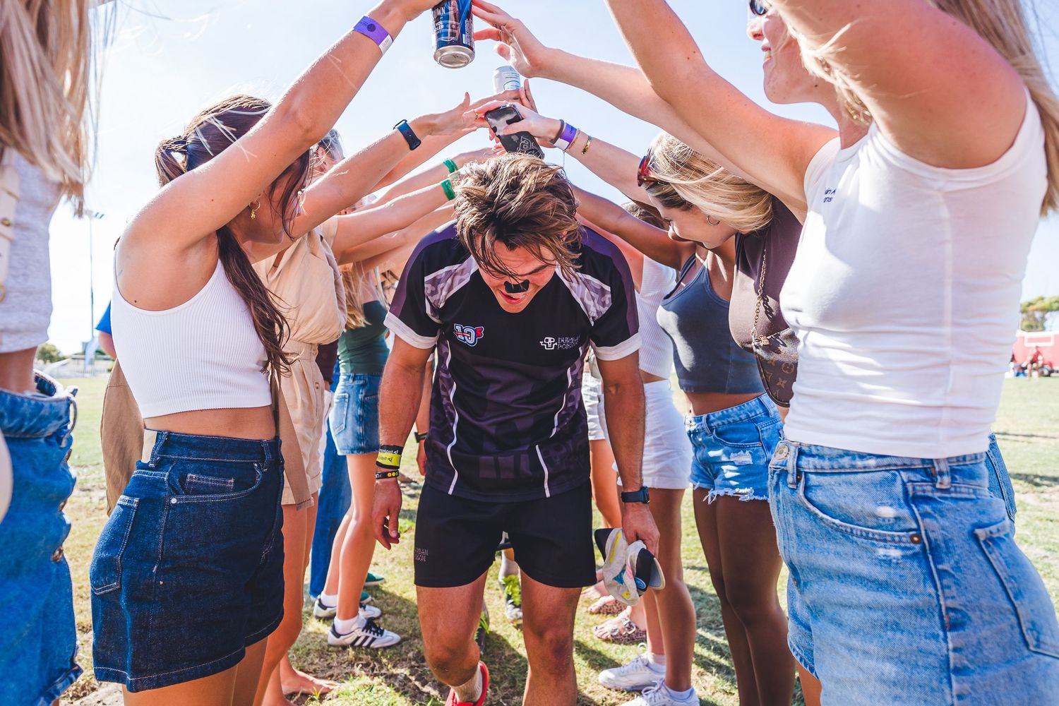 Young adults at outdoor festival raising drinks in celebration, creating a tunnel for a man in sunglasses to walk through.