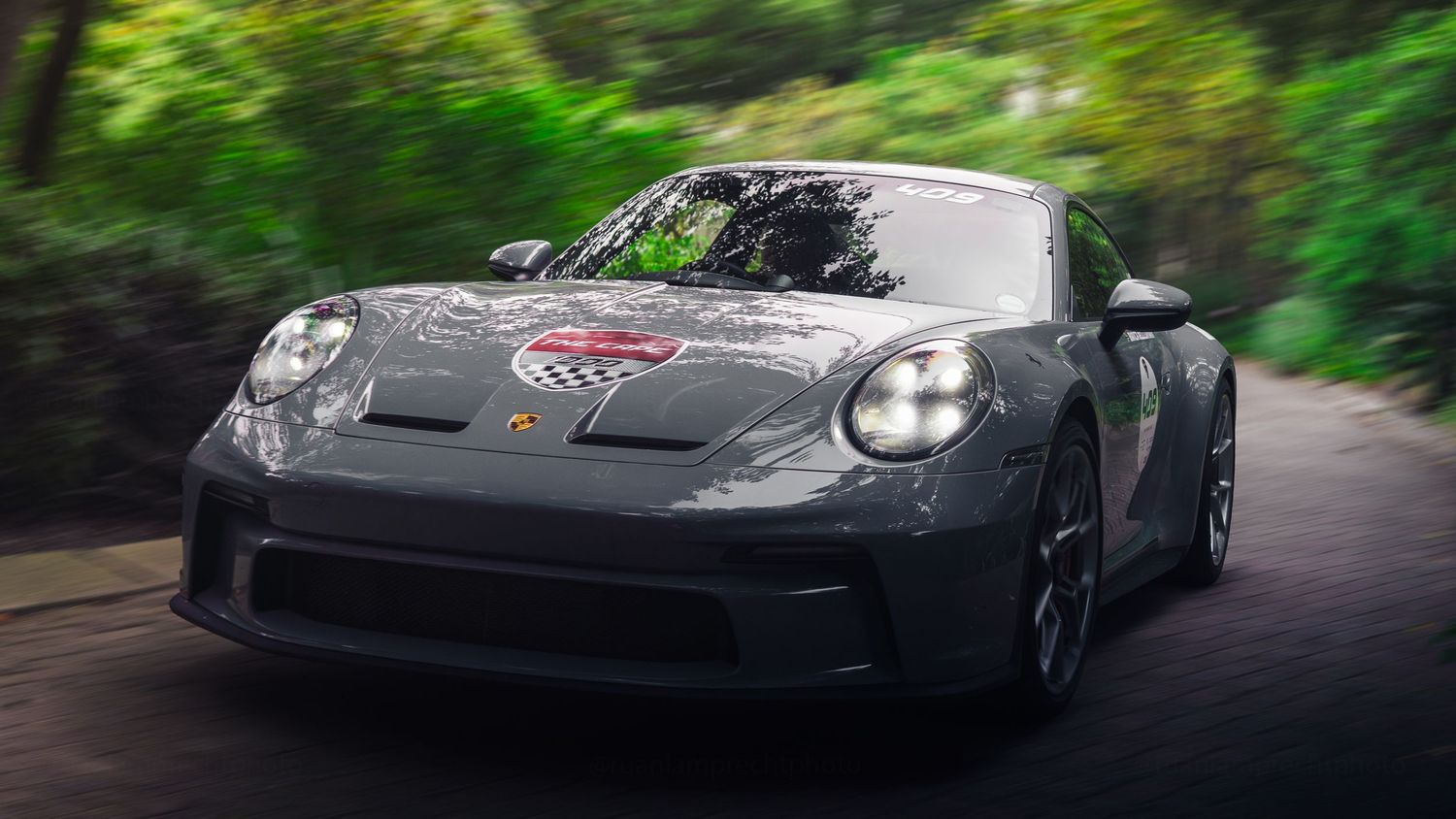 Gray Porsche 911 GT3 driving on a tree-lined road, showcasing its sleek aerodynamic design and sporty front fascia.