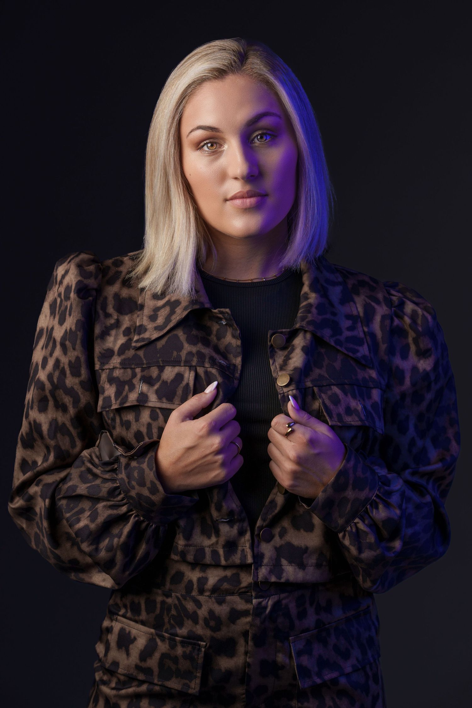 Blonde woman in leopard print jacket posing confidently against dark background with purple lighting.