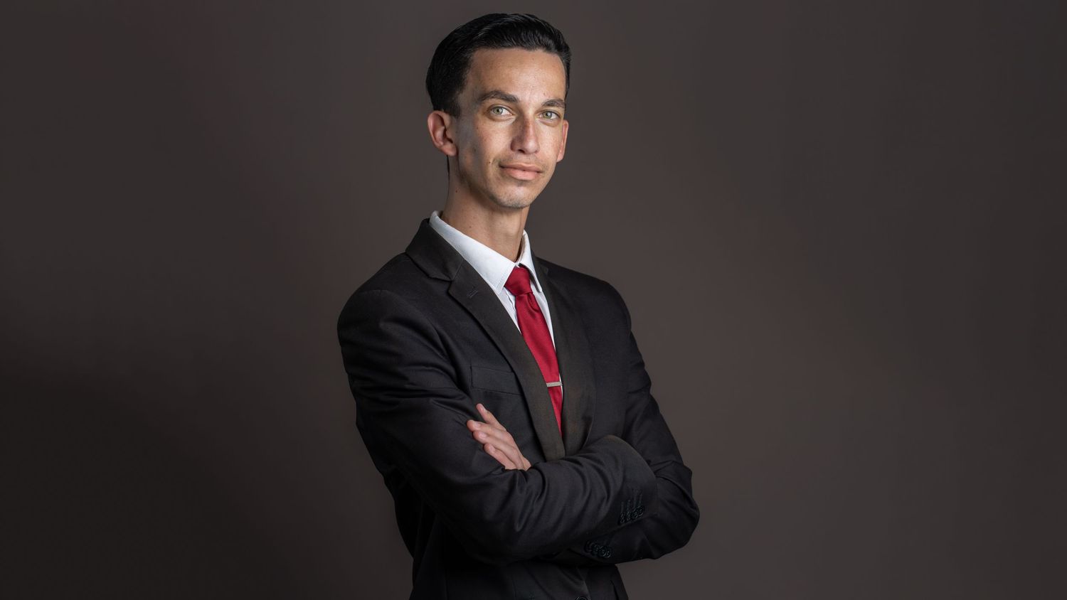 Professional headshot of man in suit and red tie against dark background.