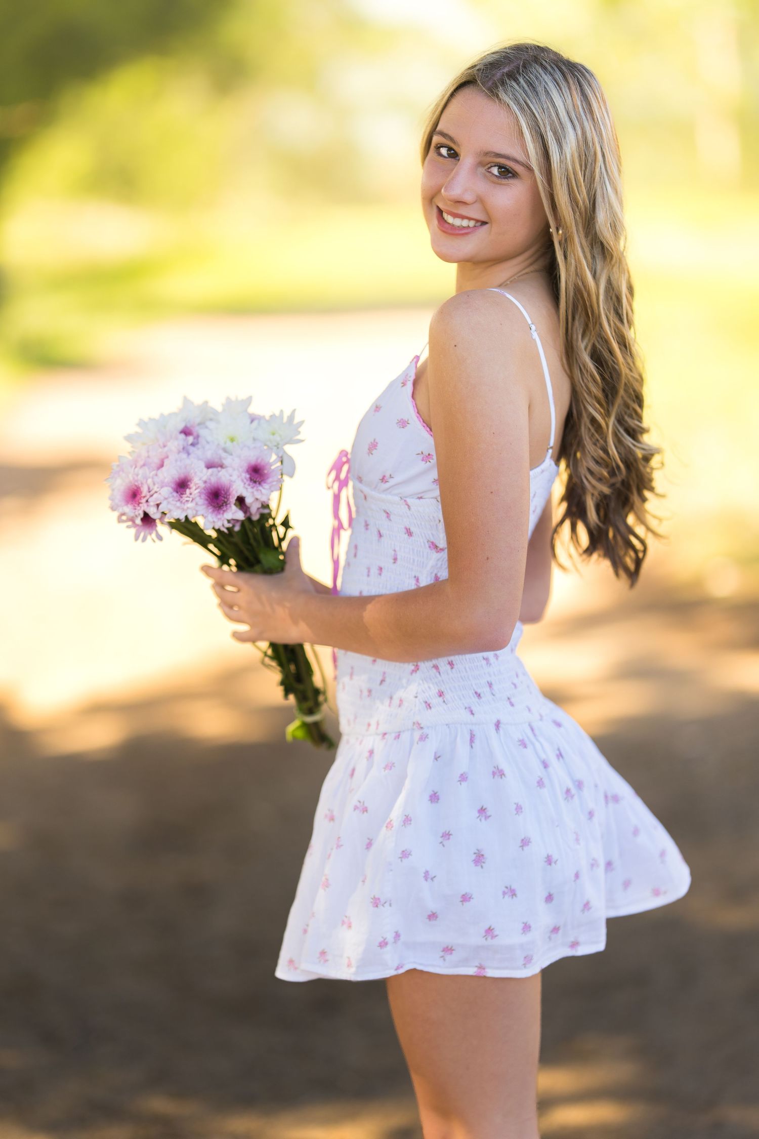A child in a white floral dress with ruffles twirls playfully in the sunlit outdoors.