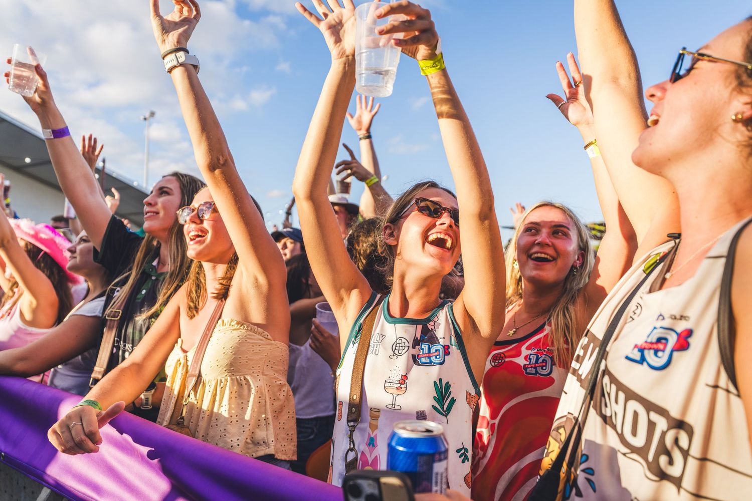 Excited crowd of young women cheering and dancing at an outdoor music festival on a sunny day.