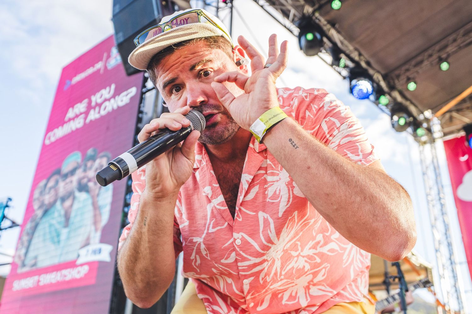 Man in pink floral shirt performing on stage with microphone at outdoor music festival.