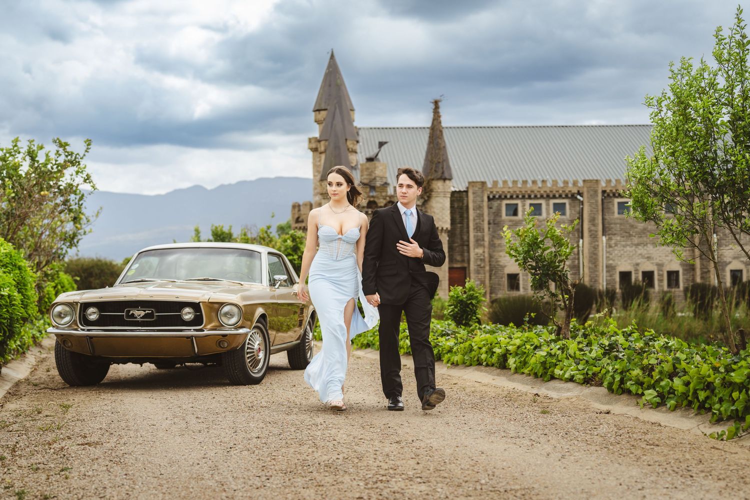 Couple in formal attire walking beside a vintage gold Ford Mustang outside a stone castle venue.
