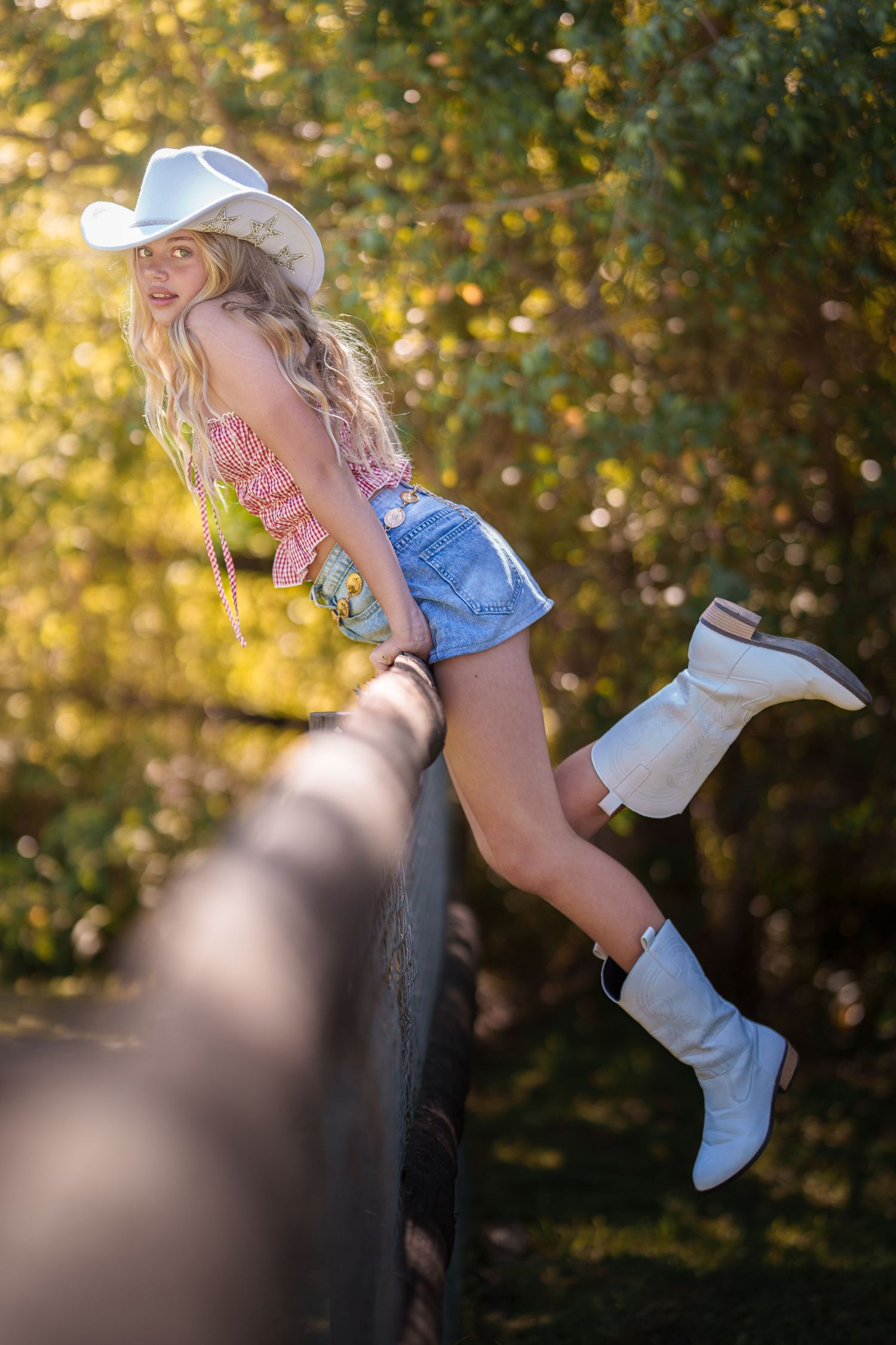 A person wearing white cowboy boots and denim shorts poses playfully on a dark fence in golden sunlight.