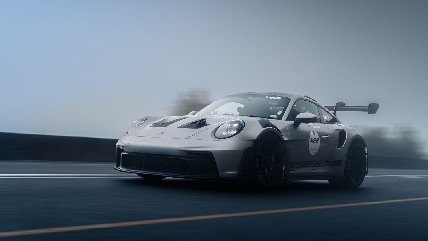 A white Porsche 911 GT3 RS race car speeds on a wet track in foggy conditions, featuring a large rear wing.