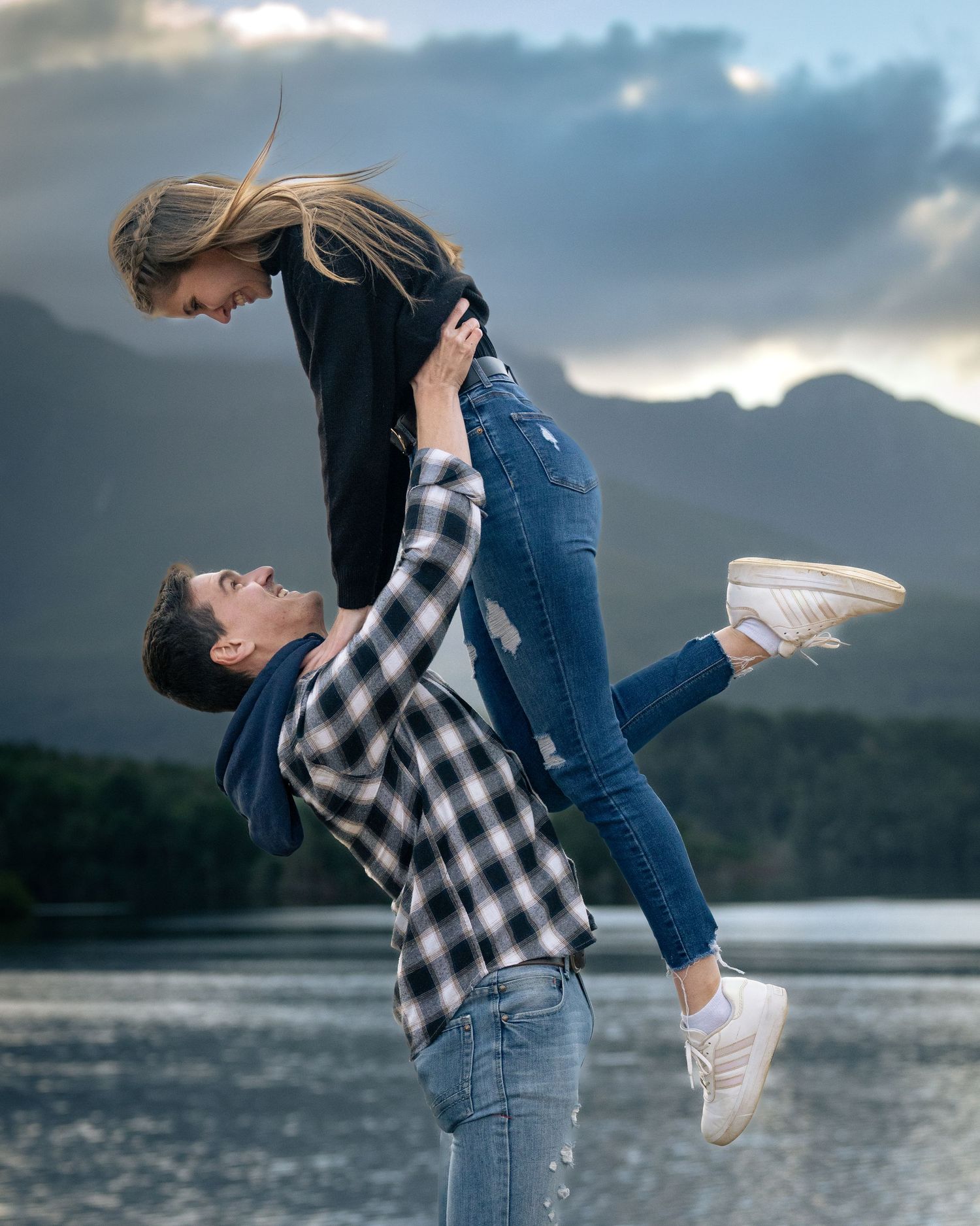 A couple embraces playfully by a lake with mountains in the background at dusk.