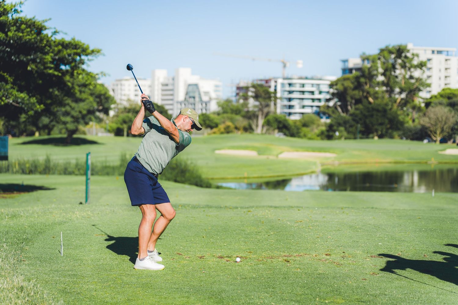 Golfer completing a swing on a lush green golf course with trees and buildings in the background on a sunny day.