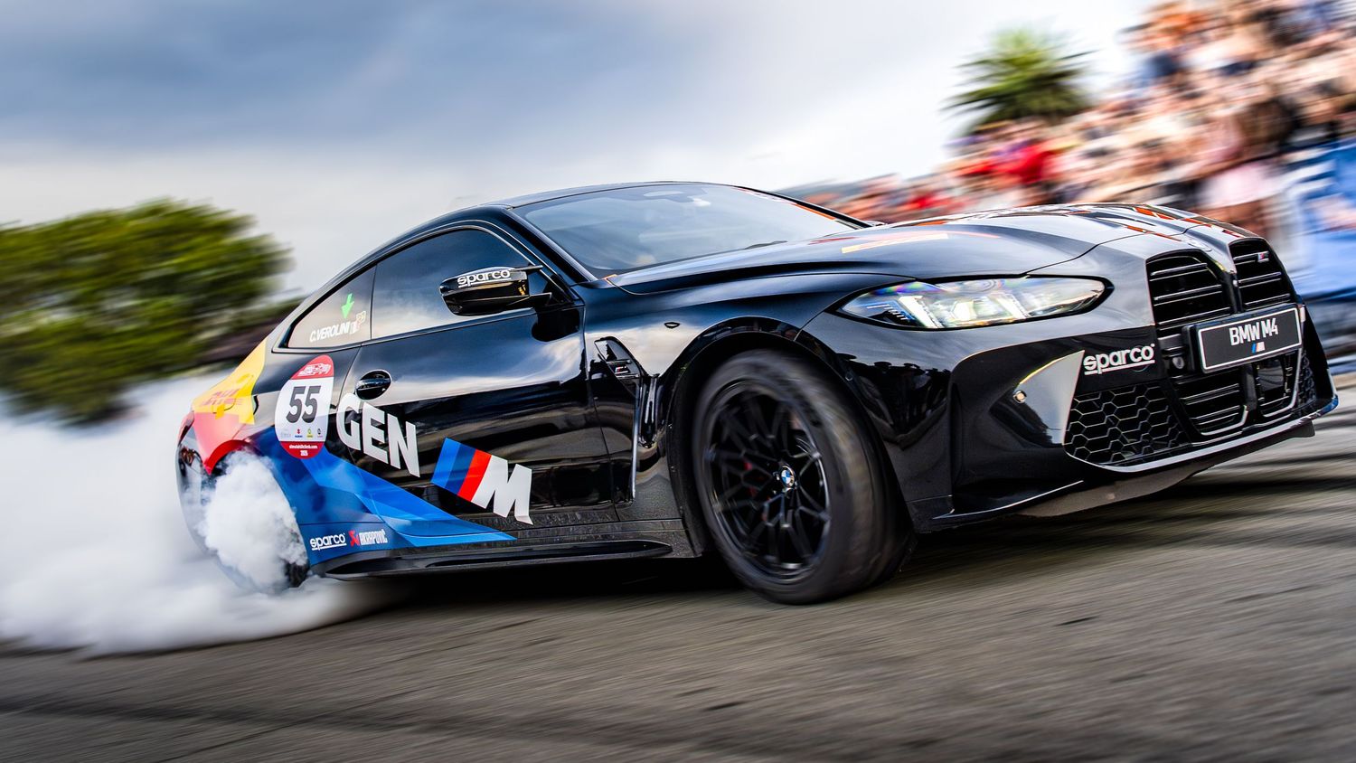 Black BMW M4 GEN drifting with smoke from tires at motorsport event.