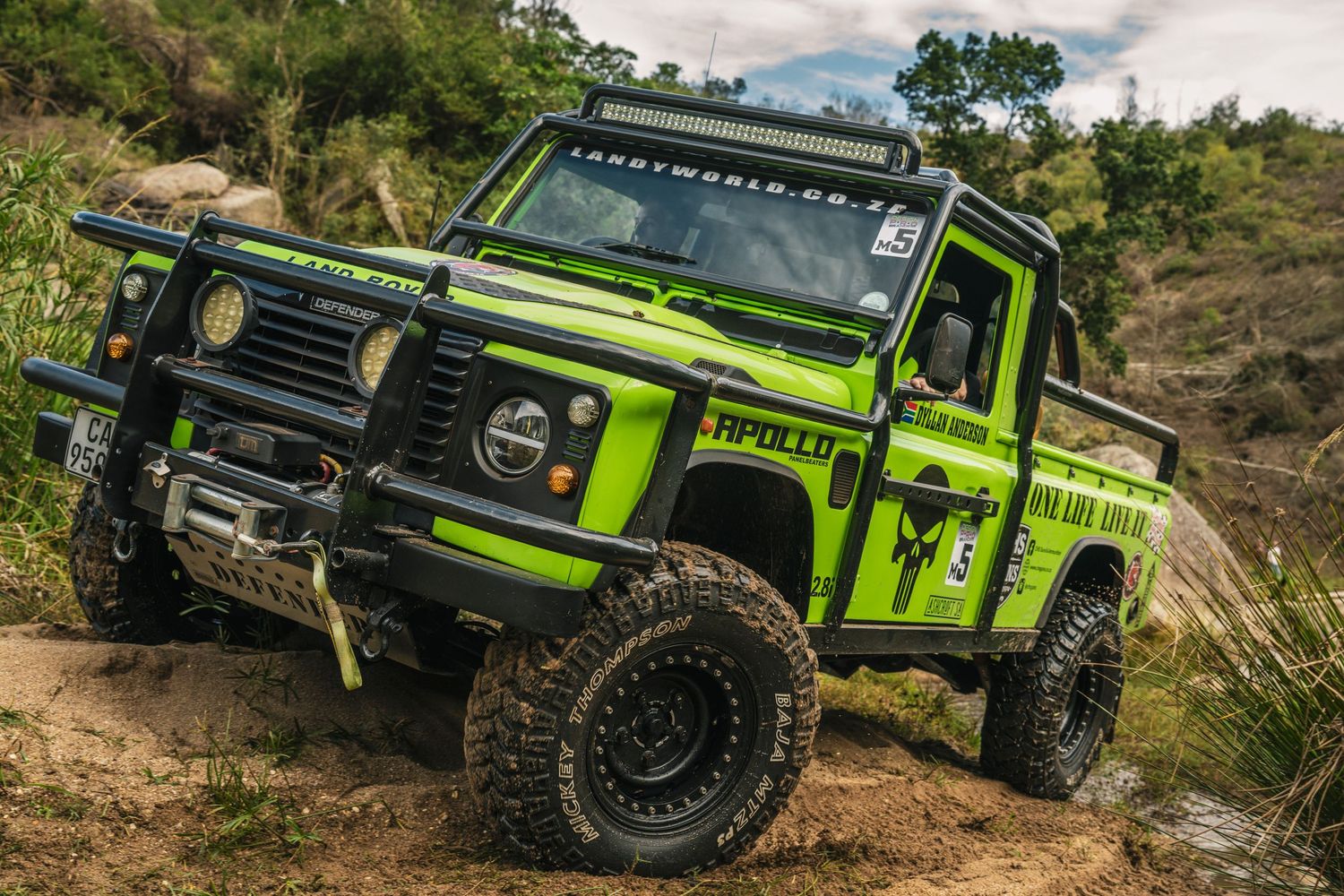 Bright green modified Land Rover Defender pickup truck with large off-road tires navigating a muddy trail outdoors.