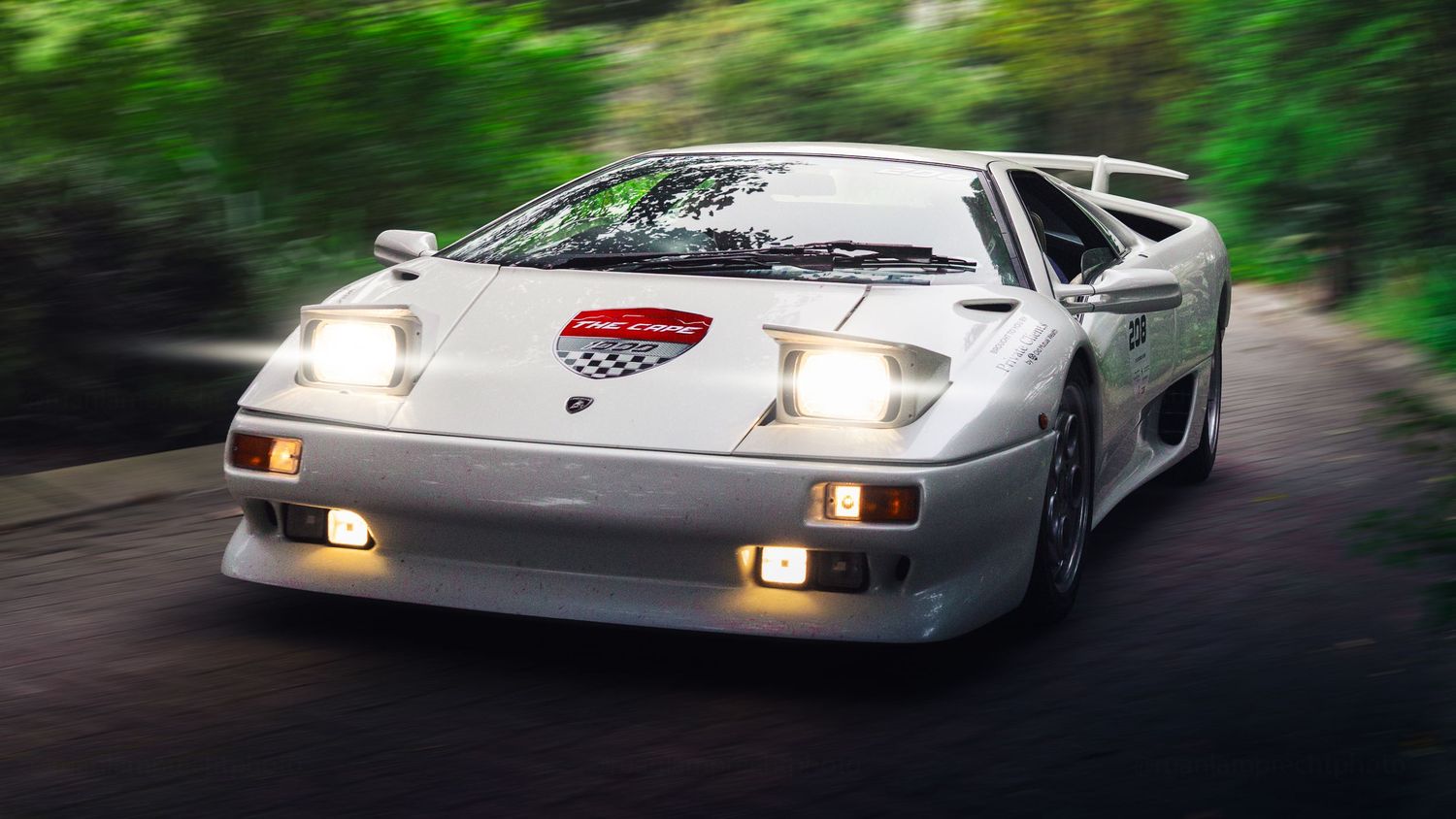 White Lamborghini Diablo supercar driving on a tree-lined road with headlights on and a racing badge on the hood.