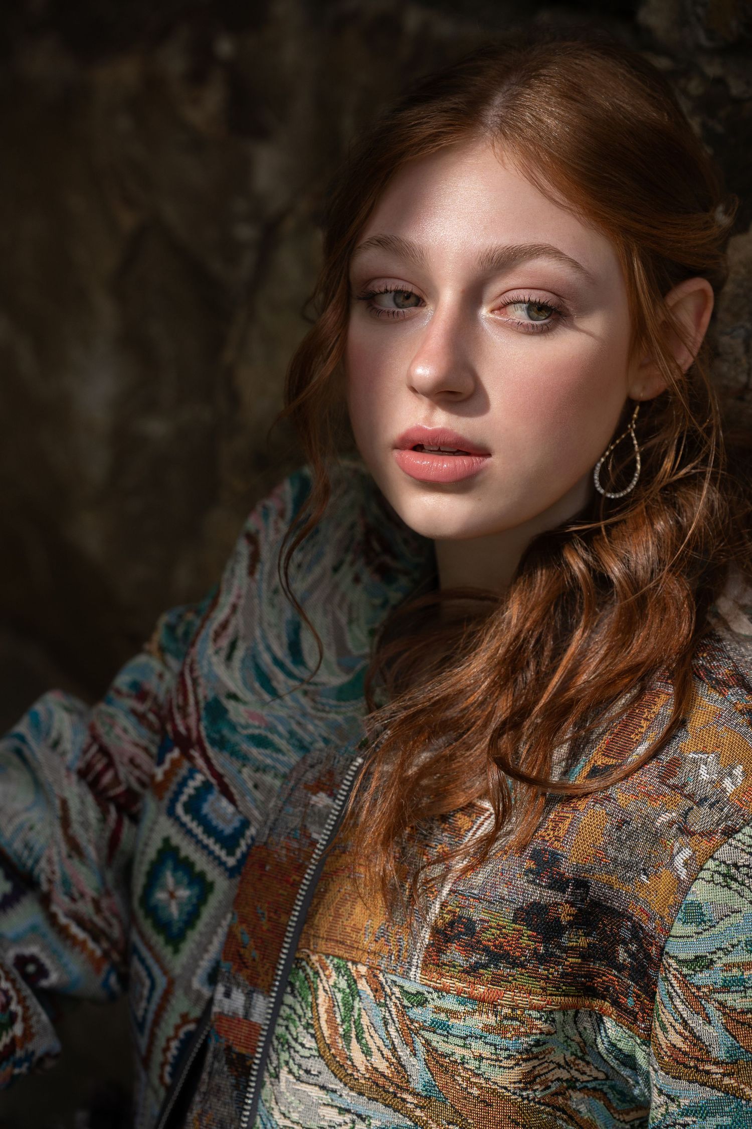 Young redhead woman in patterned jacket posing against a rocky background.