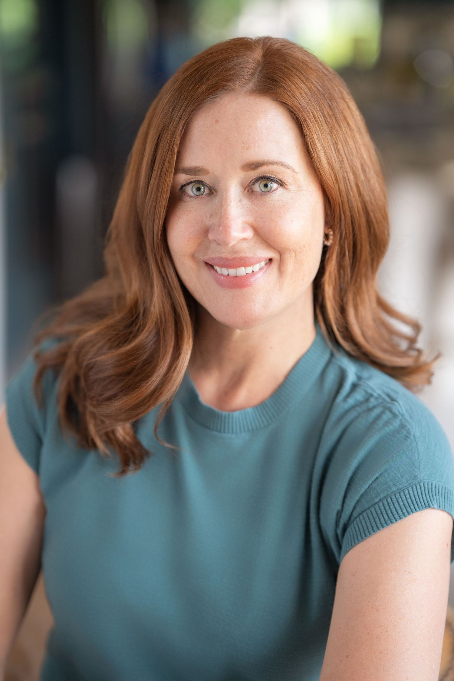 Smiling redhead woman in teal top with soft bokeh background headshot.