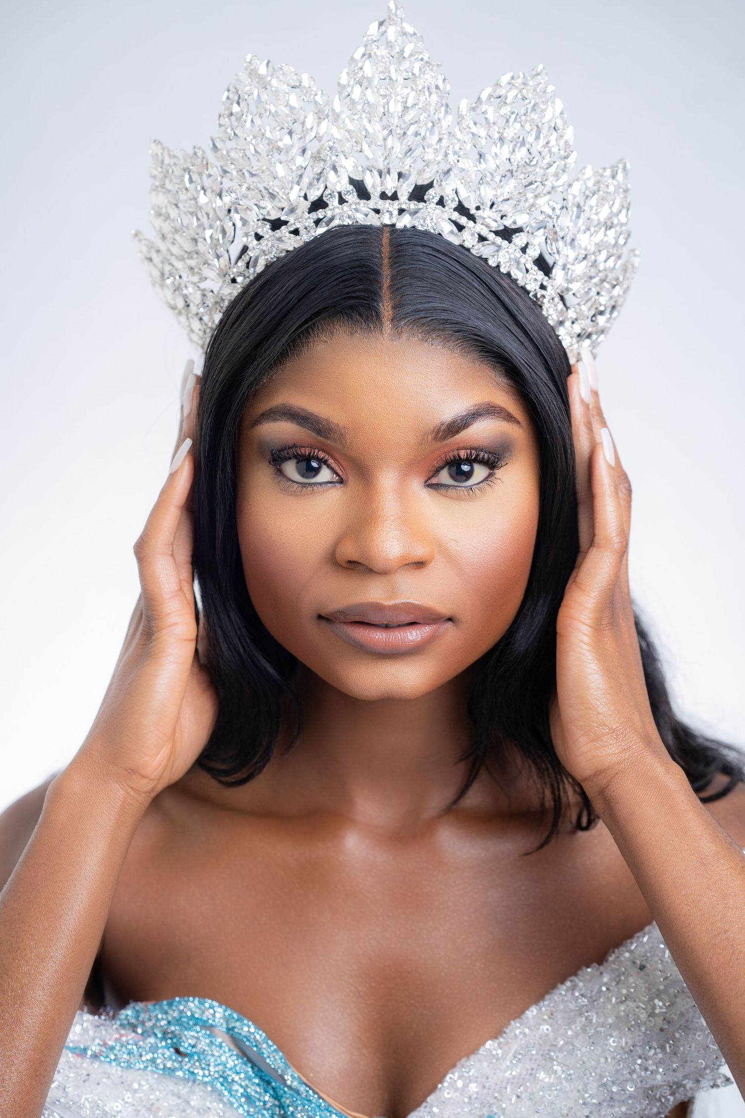 Beautiful Black woman wearing a sparkling crystal crown and strapless gown against a white background.