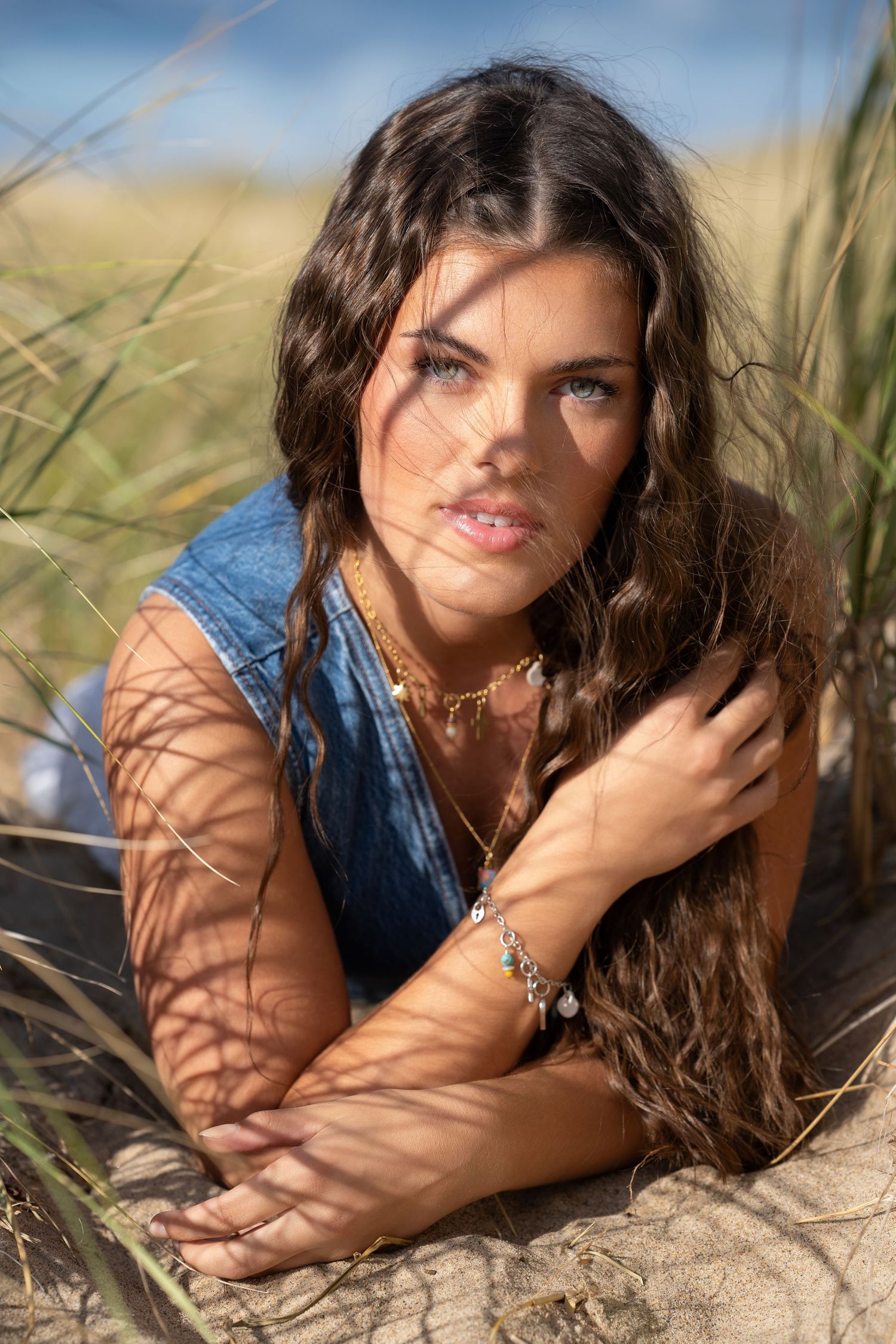 Young brunette woman posing in sand dunes wearing denim top and layered necklaces.