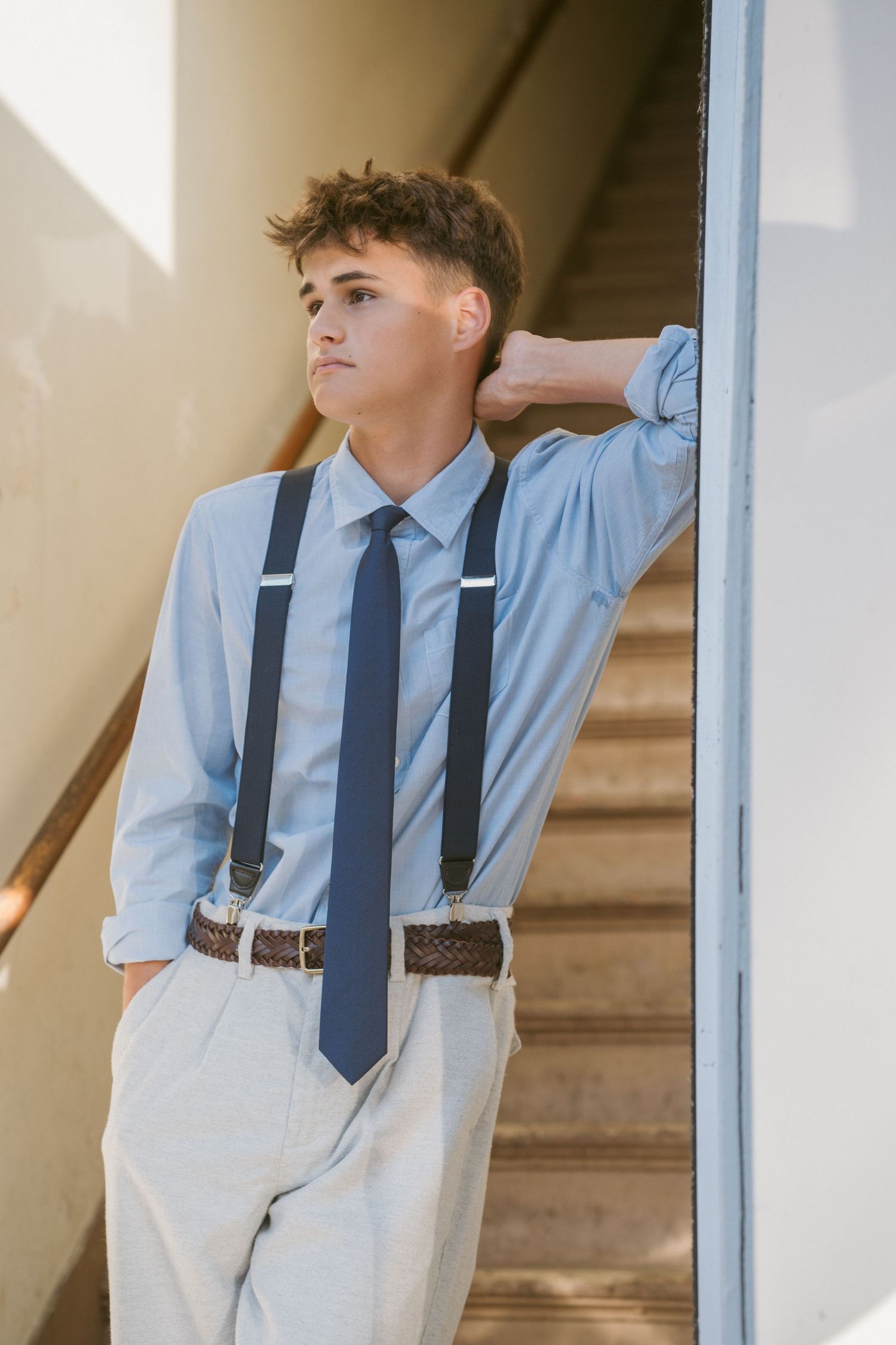 Stylish teenage boy in suspenders and tie leaning against a wall near a staircase.