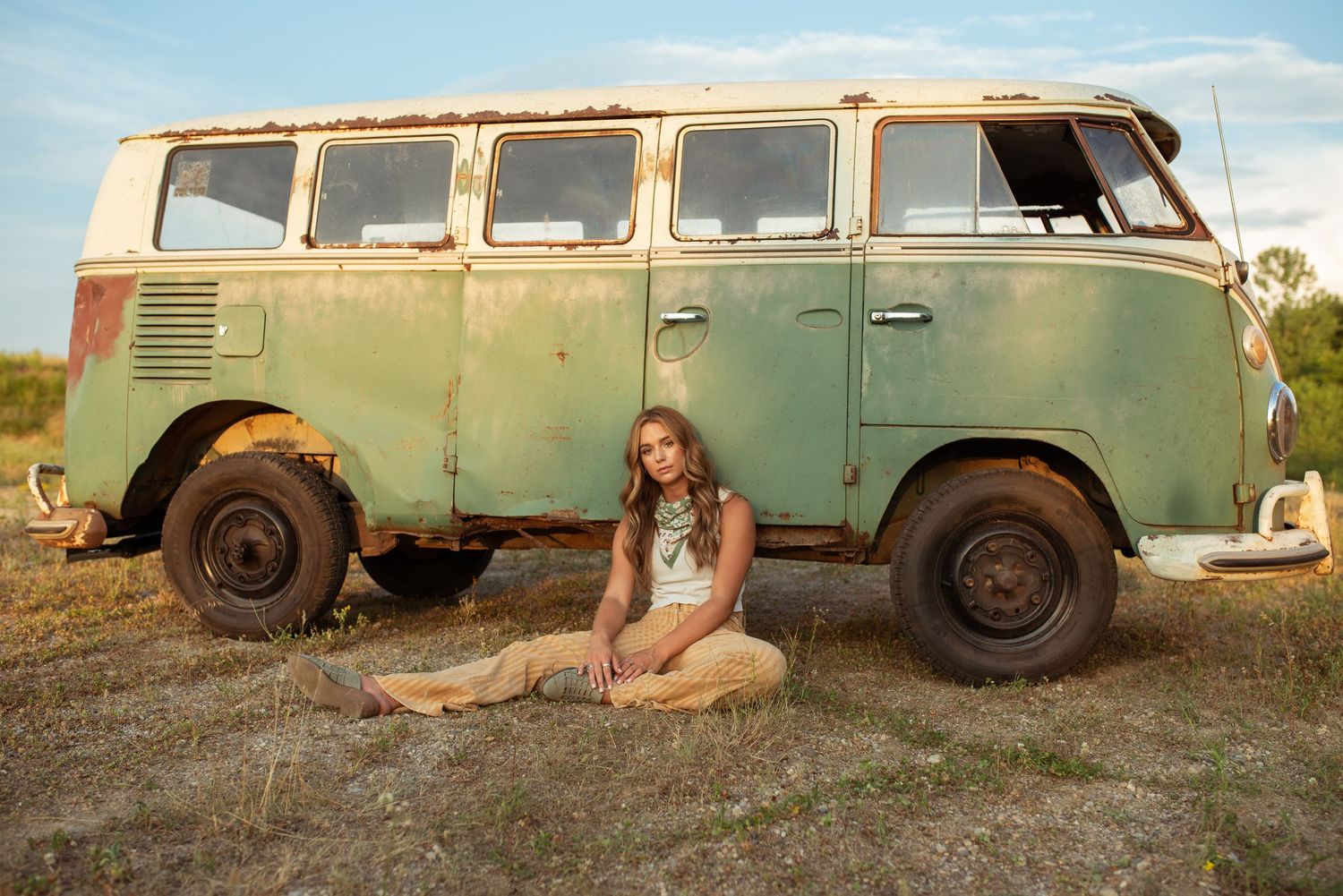 Young woman in casual outfit sitting beside a vintage rusty green Volkswagen van outdoors.